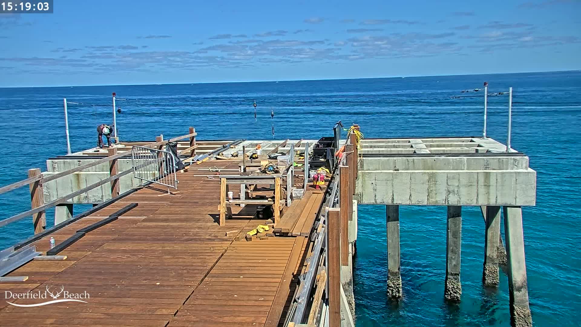 A partially constructed wooden pier extends over a calm, blue ocean under a mostly sunny sky.
