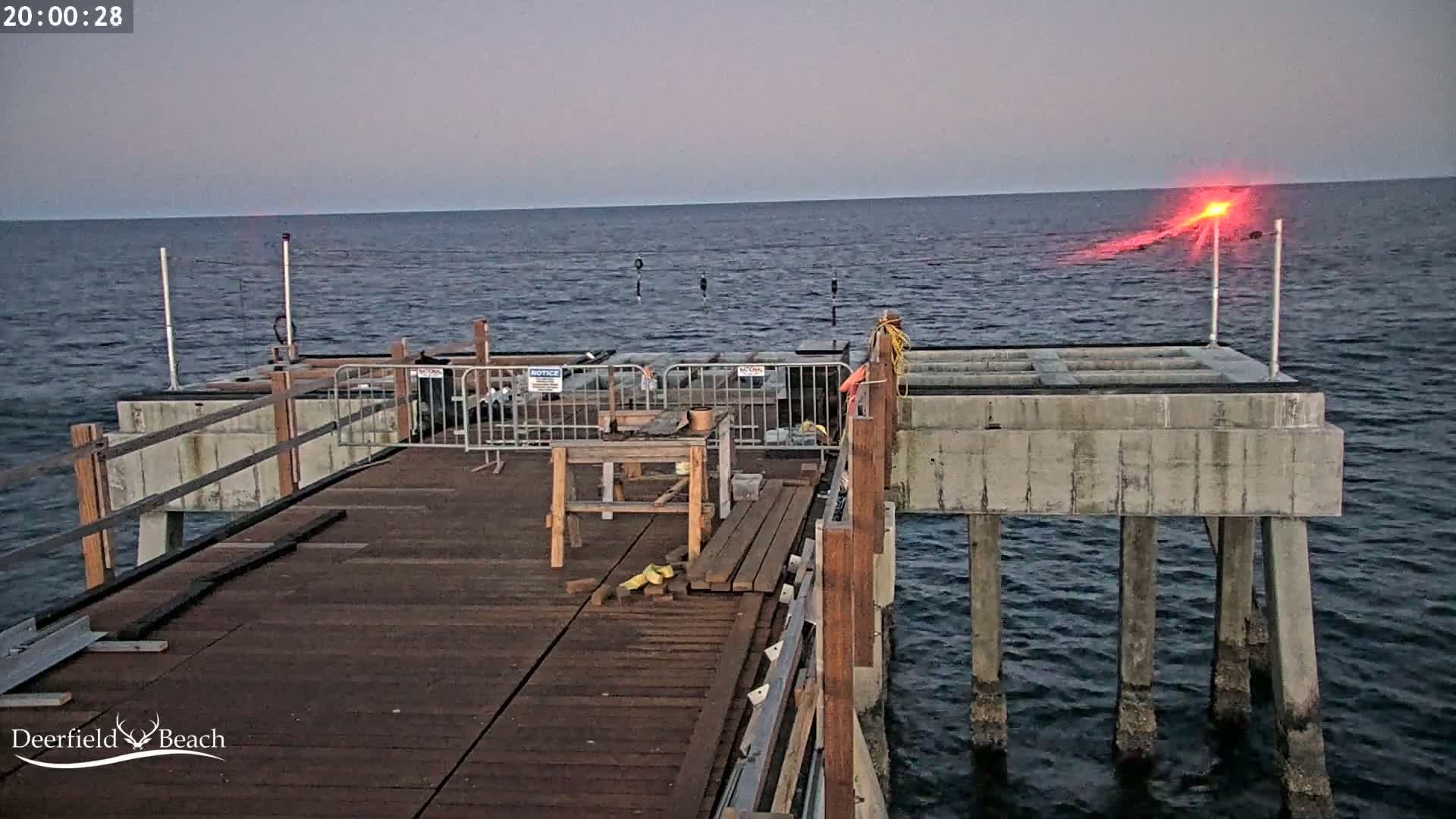 A partially constructed pier extends over dark, calm water under a twilight sky, with a red light visible on the horizon.