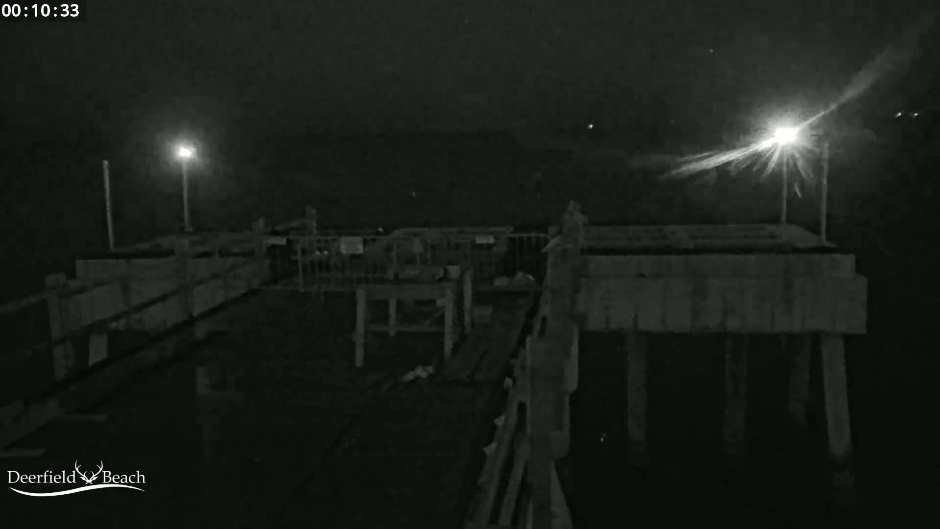 A nighttime, low-light view of a wooden pier with two illuminated lights, showing some structures and railings under dark, possibly foggy conditions.