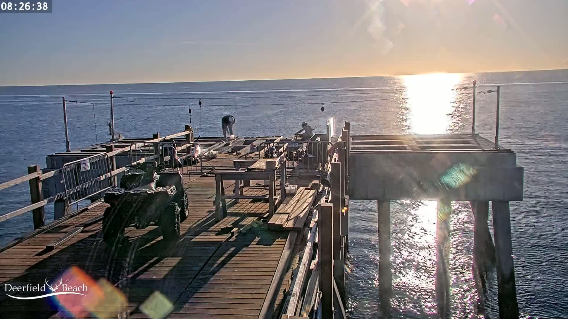 Workers are on a wooden pier over calm water, under a sunny sky at sunrise, with an ATV parked nearby.