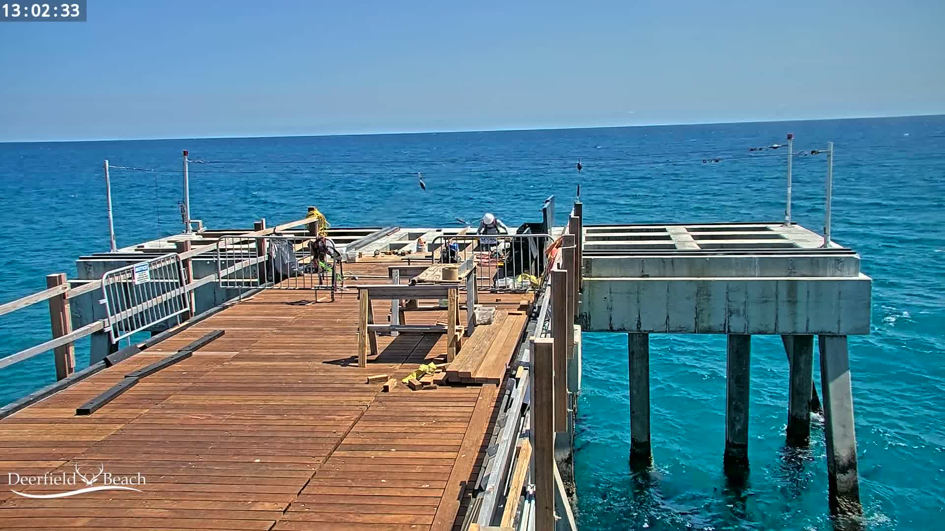 Workers are constructing a wooden deck on a pier over bright blue ocean water under a clear sunny sky.