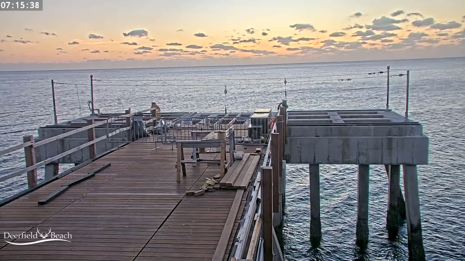 A partially constructed pier extends over a calm ocean under a sunrise sky with mostly cloudy conditions.