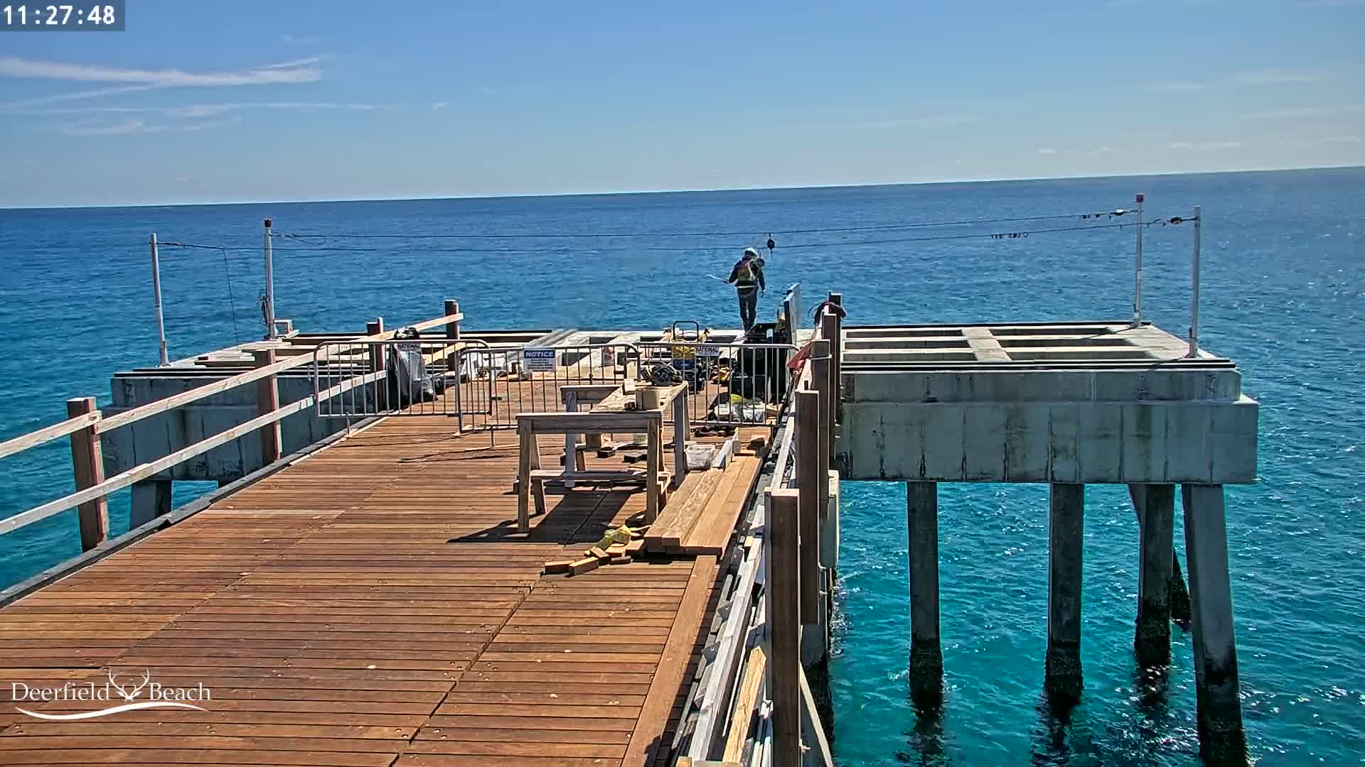 A partially-constructed wooden pier extends over a calm, blue ocean under a sunny sky, with construction workers present.