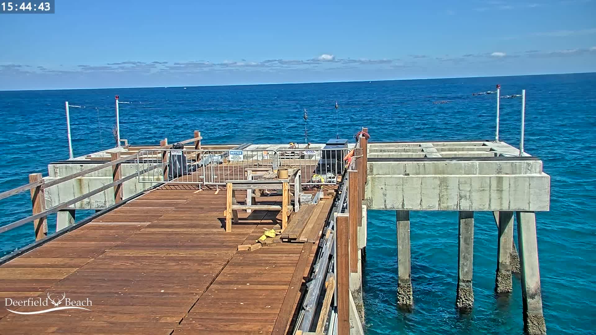 A partially constructed wooden pier extends over a calm, blue ocean under a mostly sunny sky.