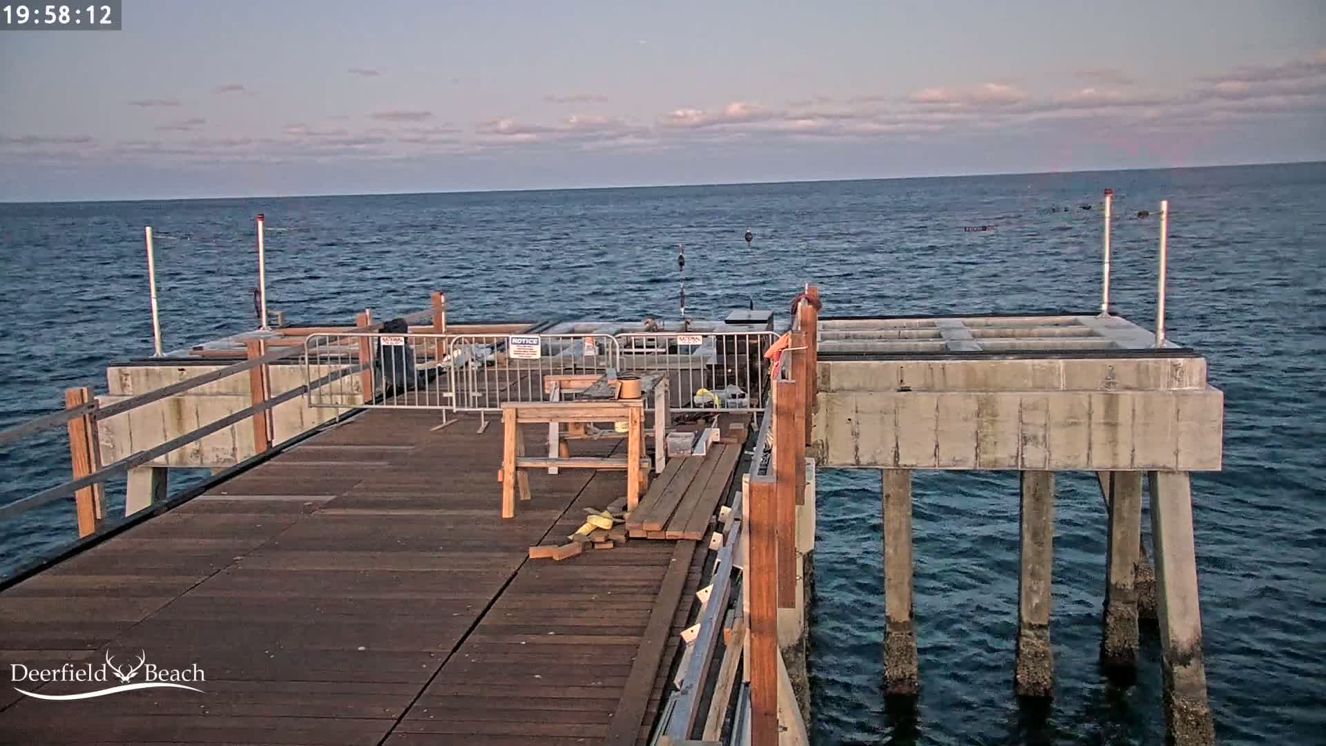 A wooden pier under construction extends over a calm, dark-blue ocean under a mostly clear, dusky sky.