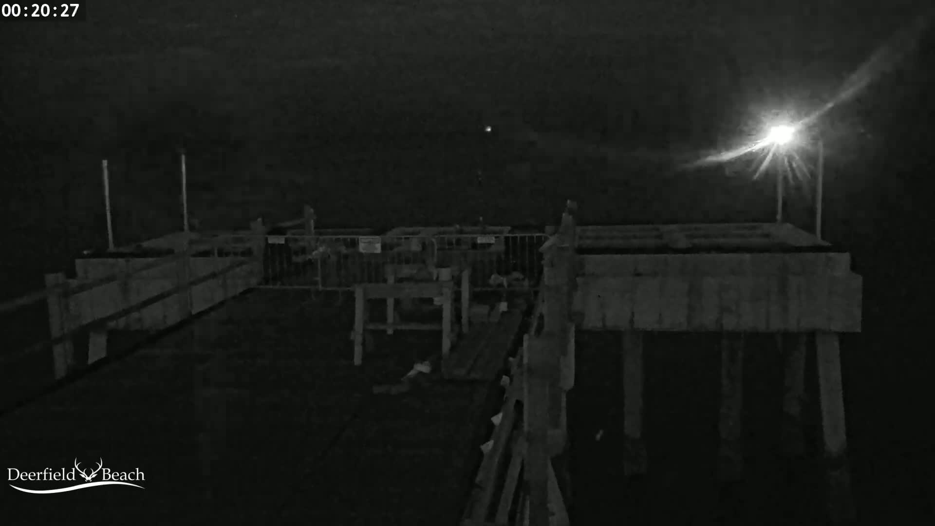 A nighttime, low-light image shows a section of a pier with wooden railings and structures under a dark sky illuminated by a single bright light.