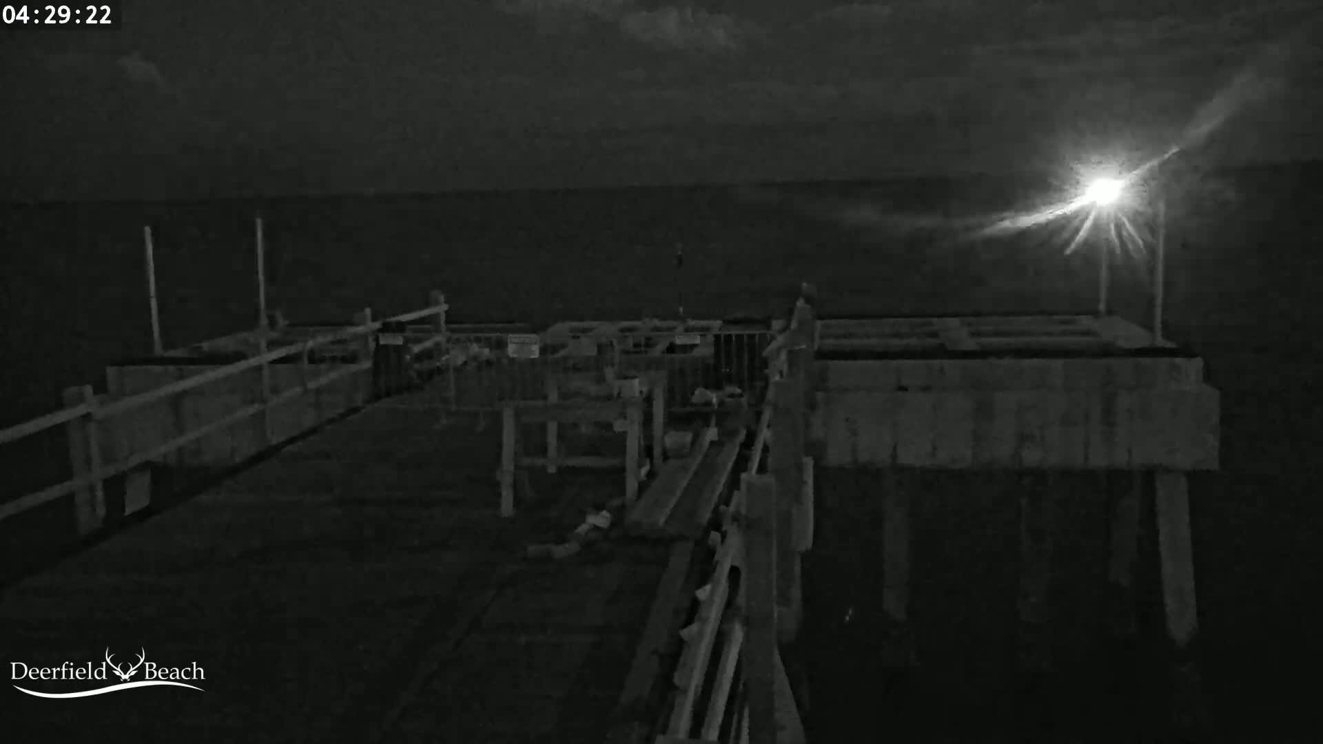 A nighttime, low-light image shows a pier extending over dark water under a partly cloudy sky, illuminated by a bright light source.