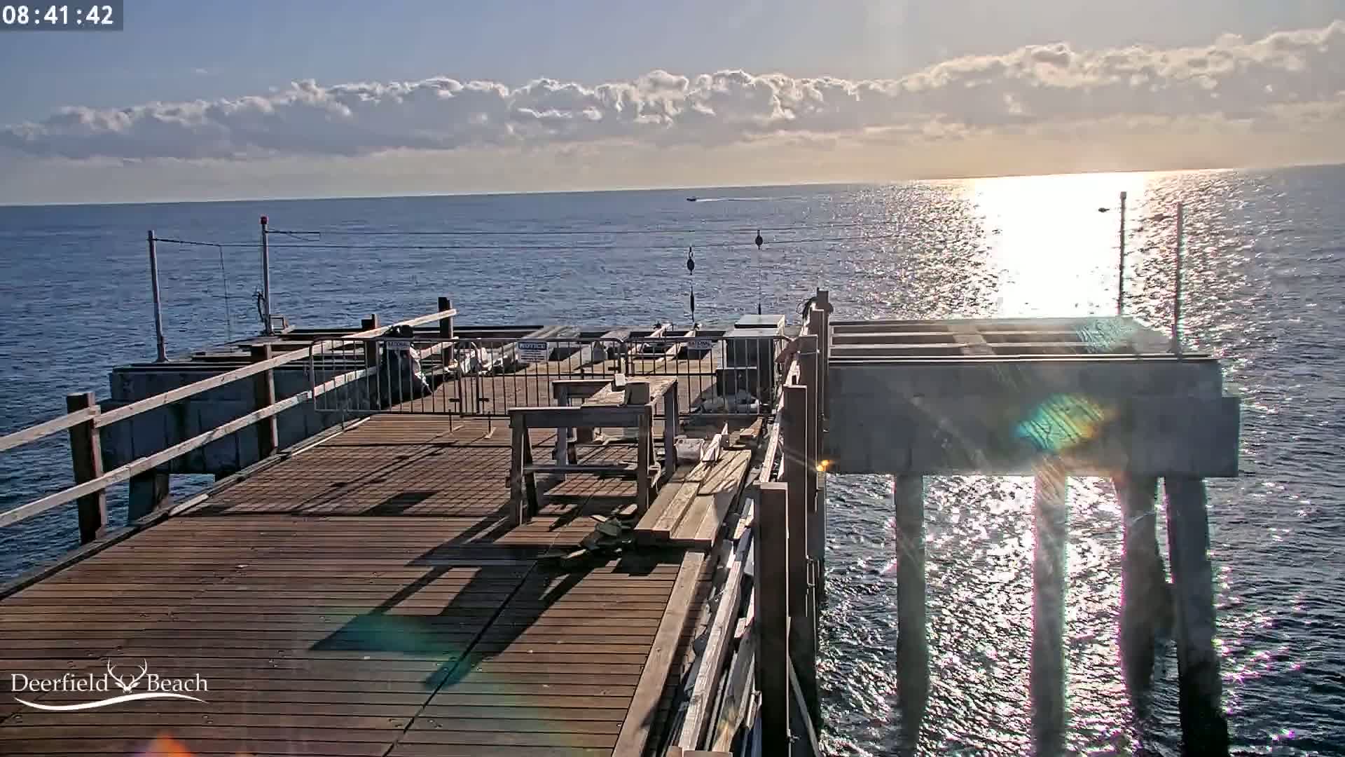 A wooden pier extends over a calm, sunlit ocean under a partly cloudy sky.