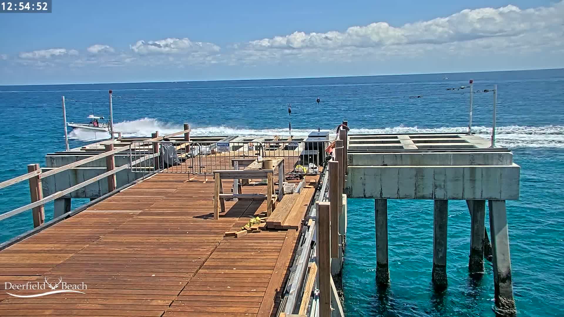 A partially constructed wooden pier extends over a blue ocean under a partly cloudy sky, with a boat speeding away in the distance.