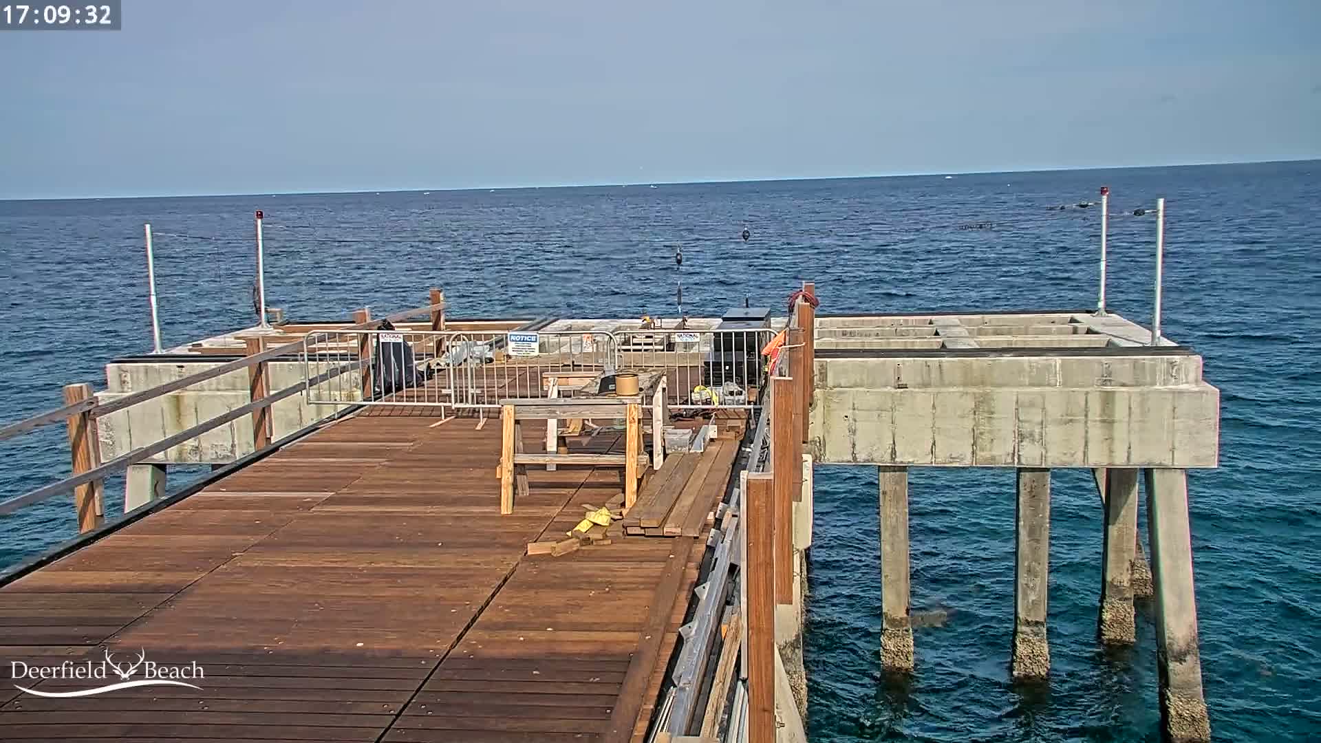 A partially-constructed wooden pier extends over a calm, blue ocean under a clear sky.