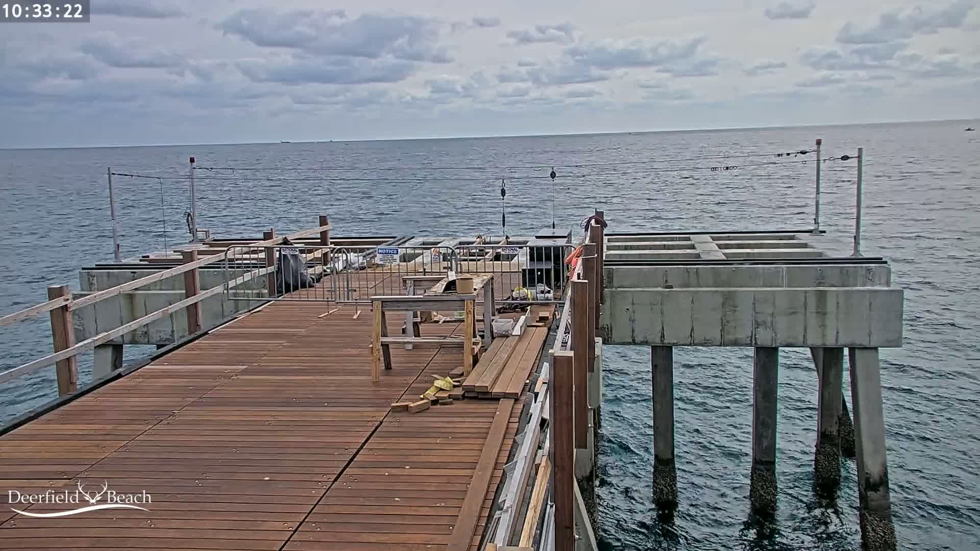 A partially constructed wooden pier extends over a calm, gray-sky ocean.