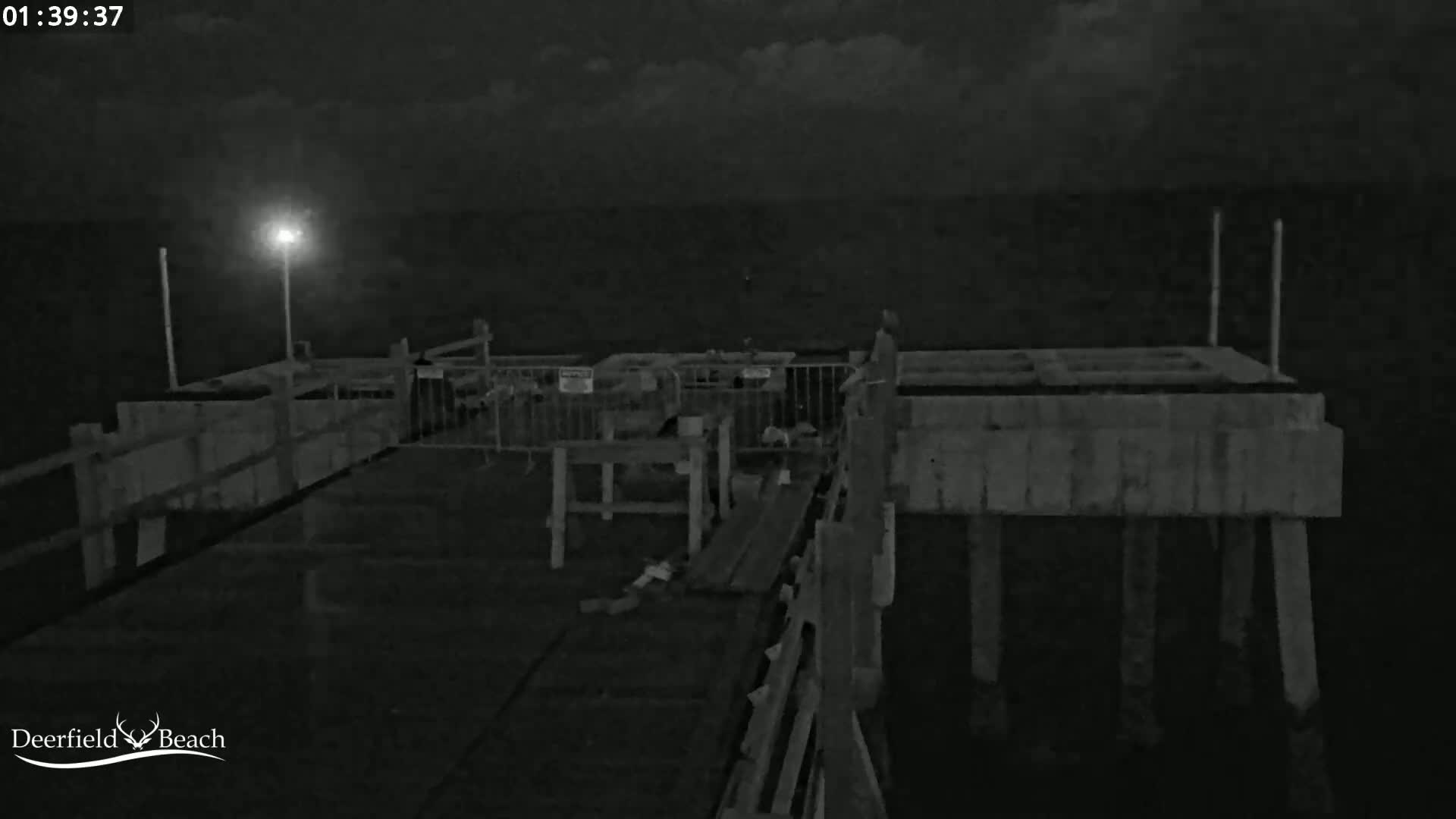 A nighttime, low-light image shows a section of a pier over dark water, illuminated by a single light.