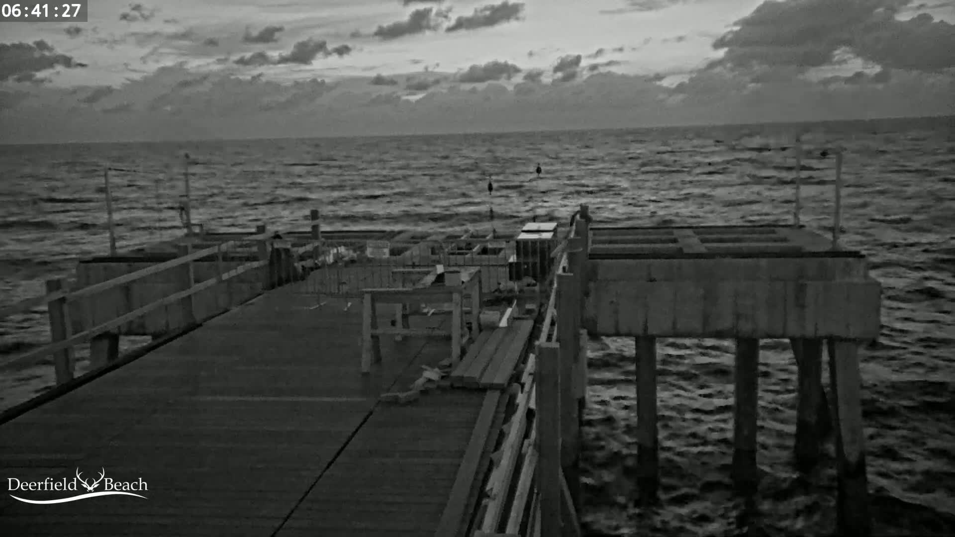 A mostly cloudy, dark pre-dawn scene shows a wooden pier extending over a choppy sea.