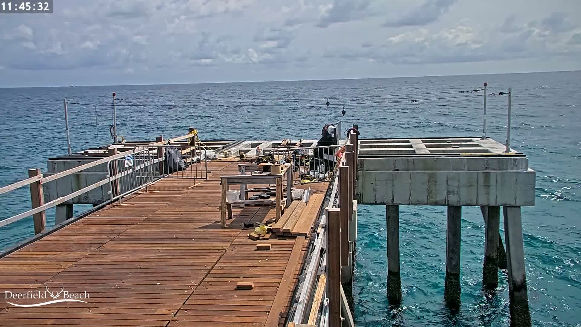 A partially constructed wooden pier extends over a blue ocean under a partly cloudy sky.