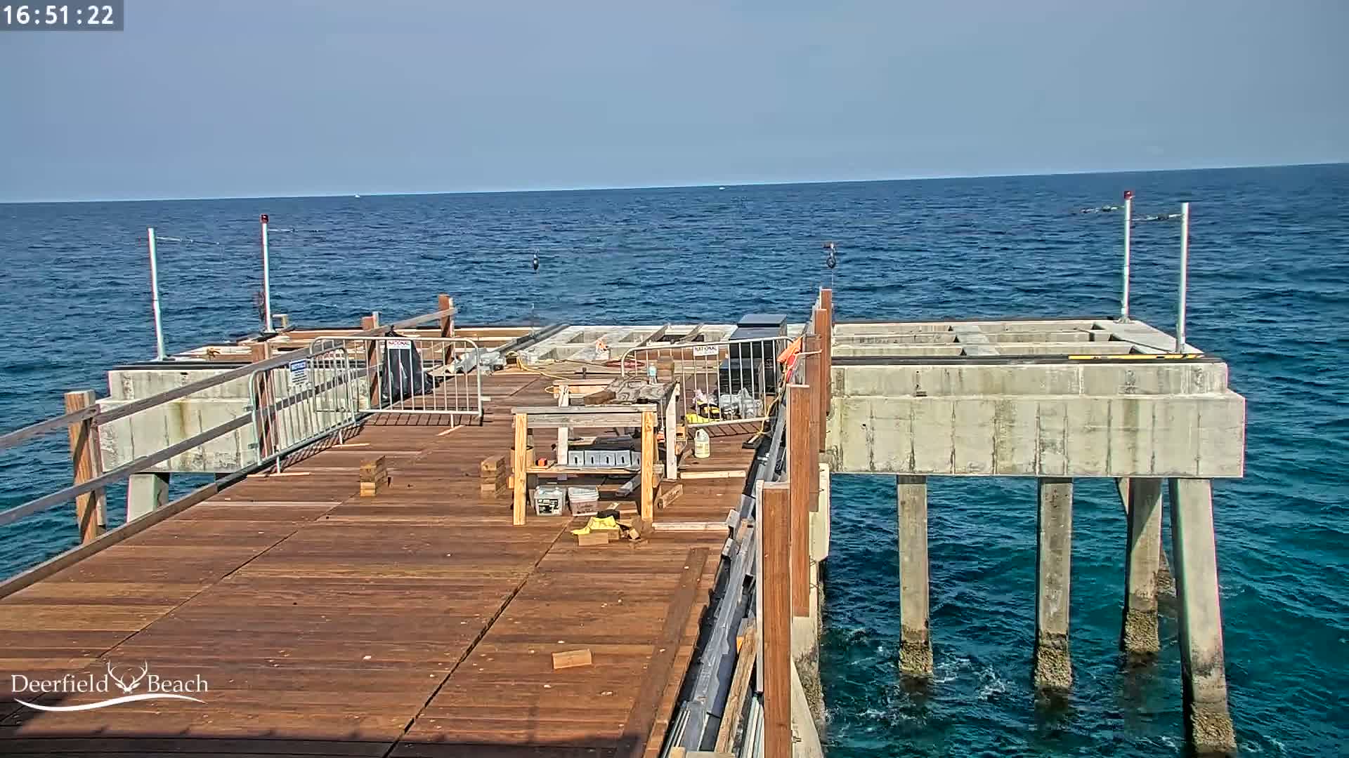 A partially-constructed wooden pier extends over a calm blue ocean under a clear sky.