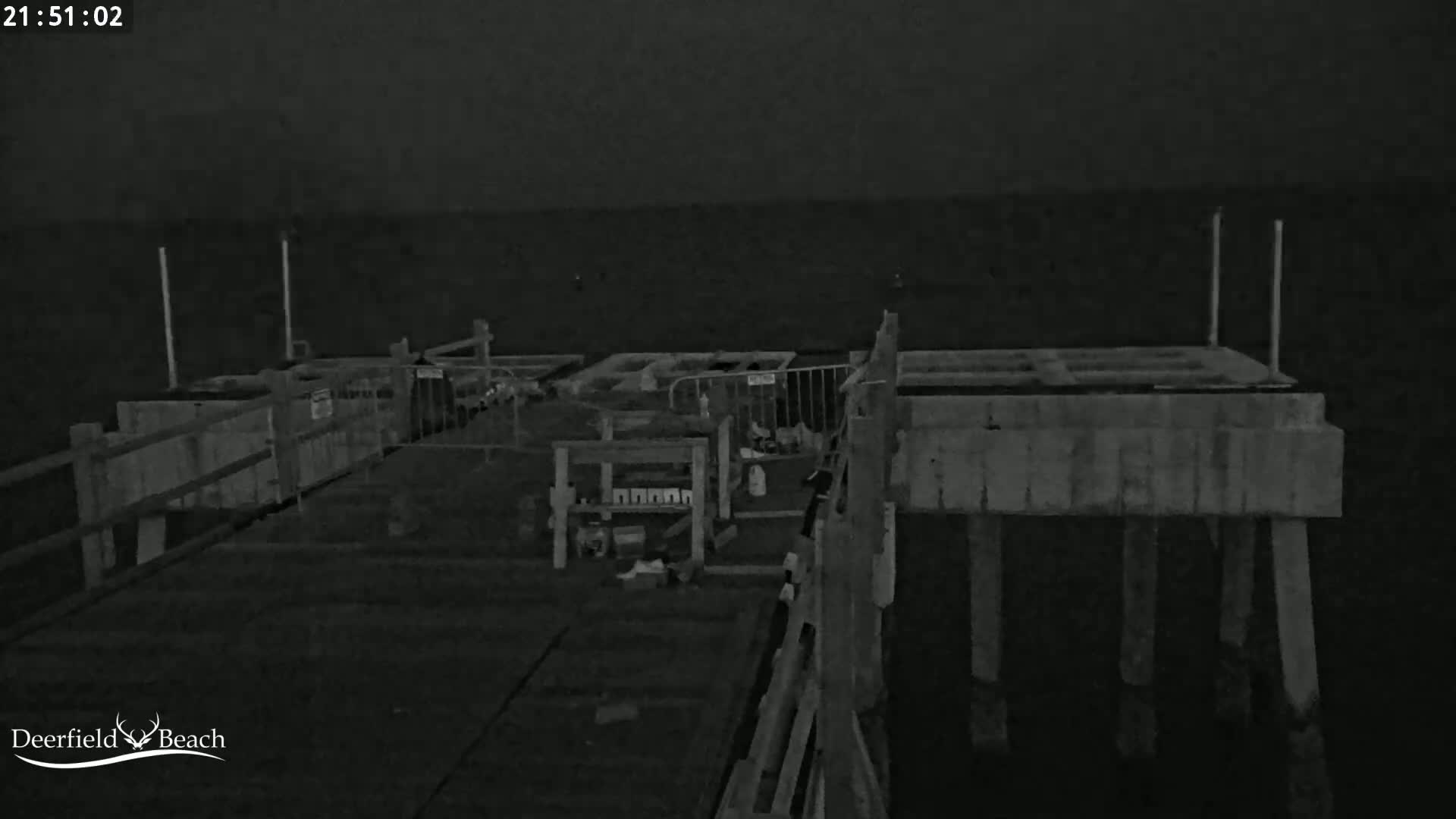 A nighttime, low-light image shows a wooden pier extending over dark water, with construction materials and barriers visible on the pier.