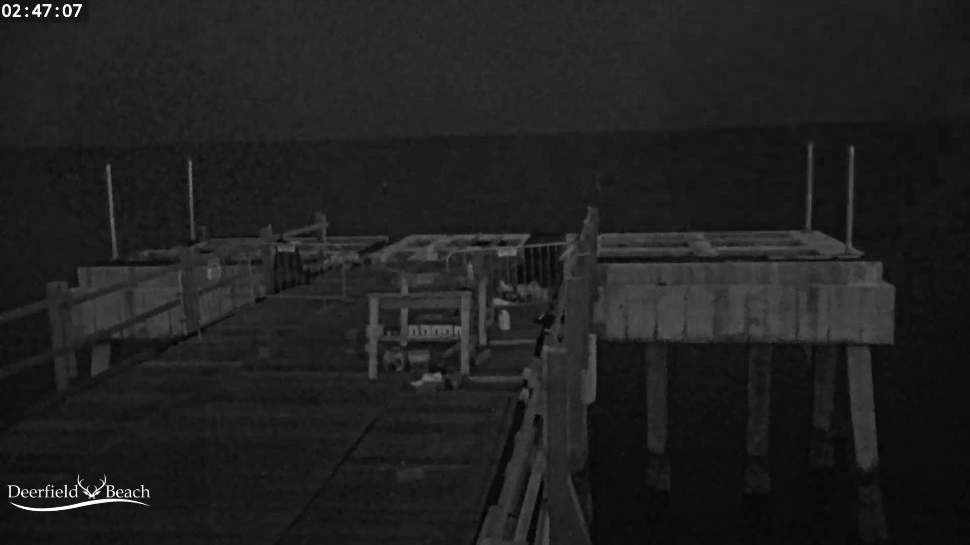 A nighttime, low-light image shows a wooden pier extending over dark water, with various structures and objects on the pier.