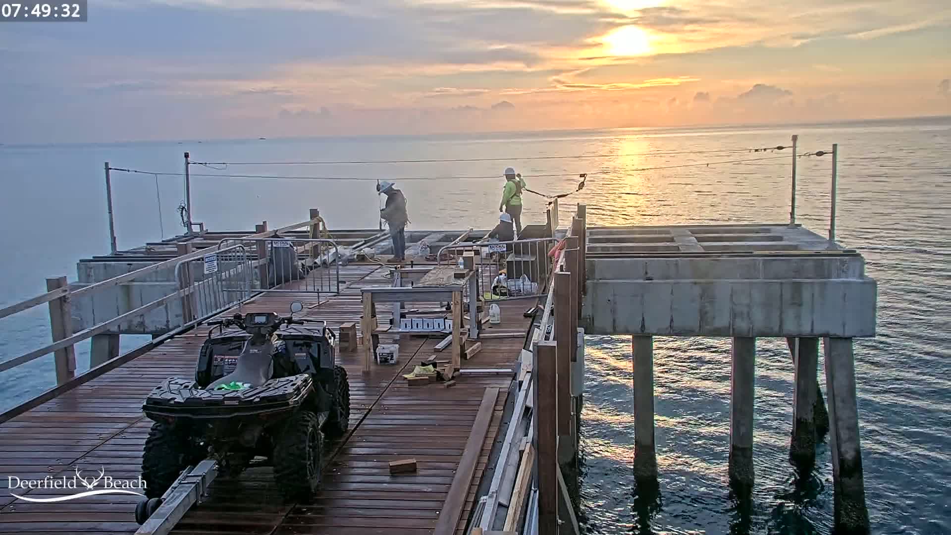 Construction workers are working on a pier during a sunrise over calm water.