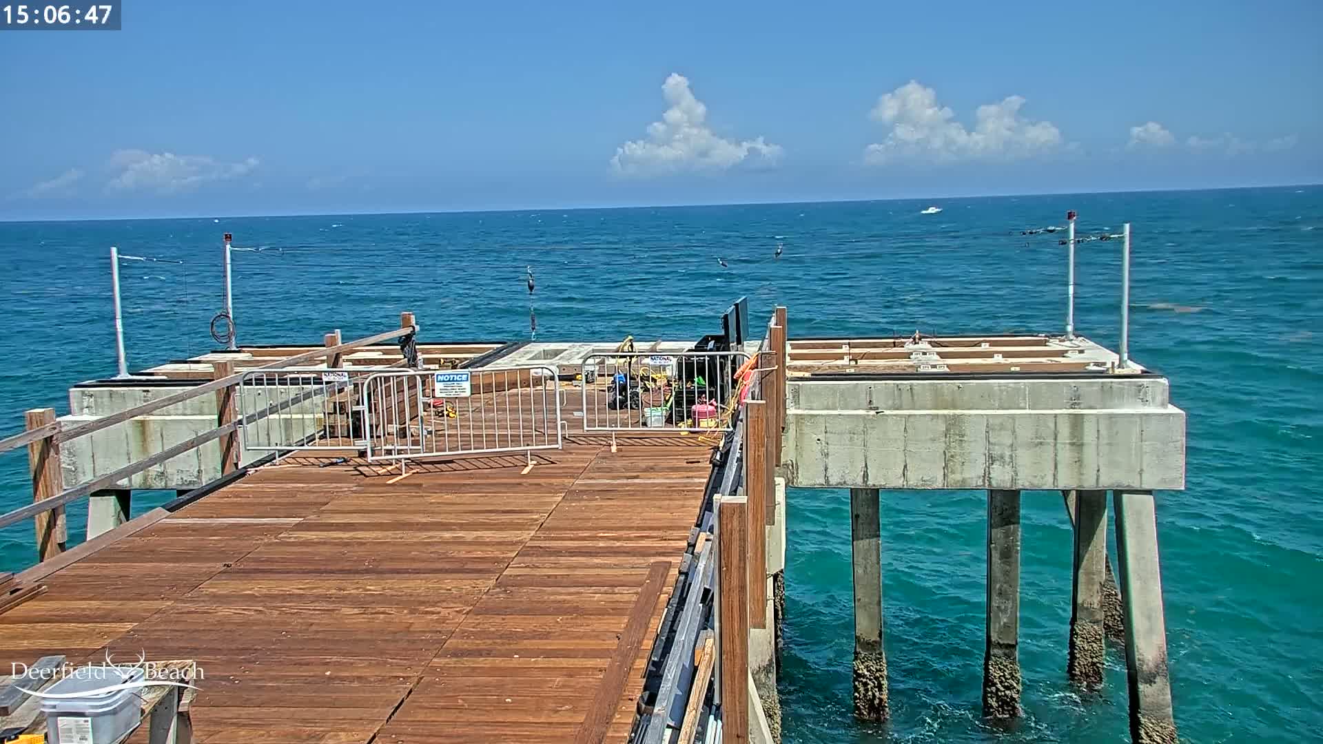 A partly constructed wooden pier extends over a teal ocean under a mostly sunny sky with a few fluffy clouds.