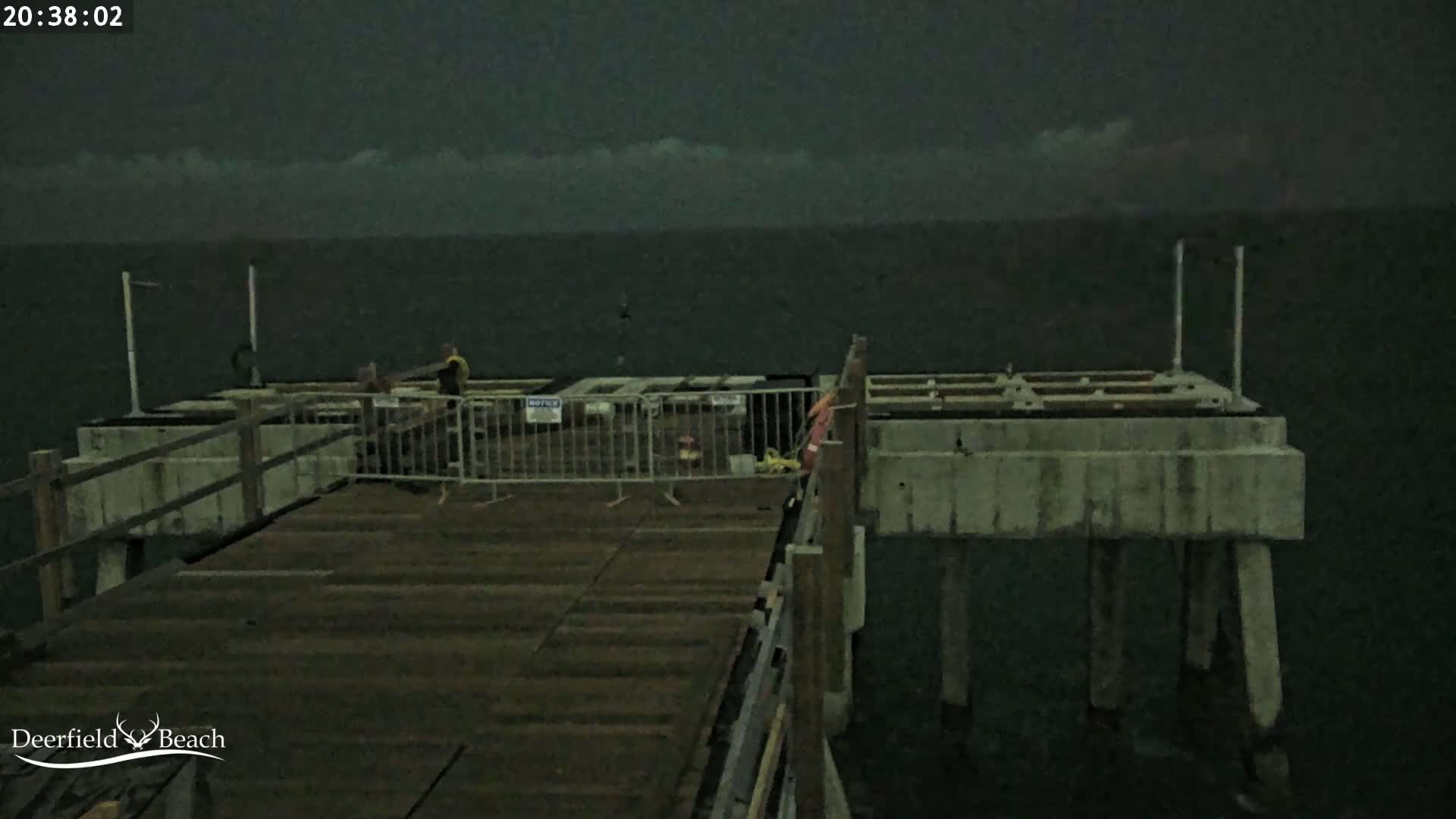 A dark, overcast night scene shows a wooden pier extending over dark water; a section of the pier is under construction, enclosed by a temporary fence, with a couple of people standing nearby.