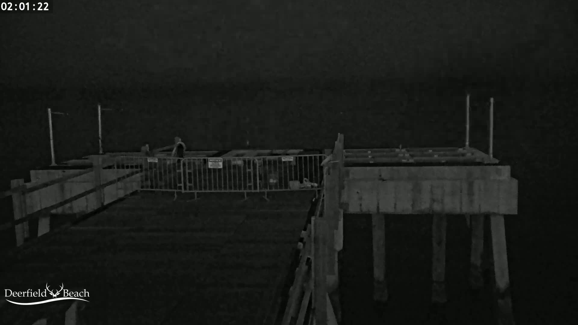A nighttime, low-light image shows a section of a pier with temporary fencing and a few indistinct figures.