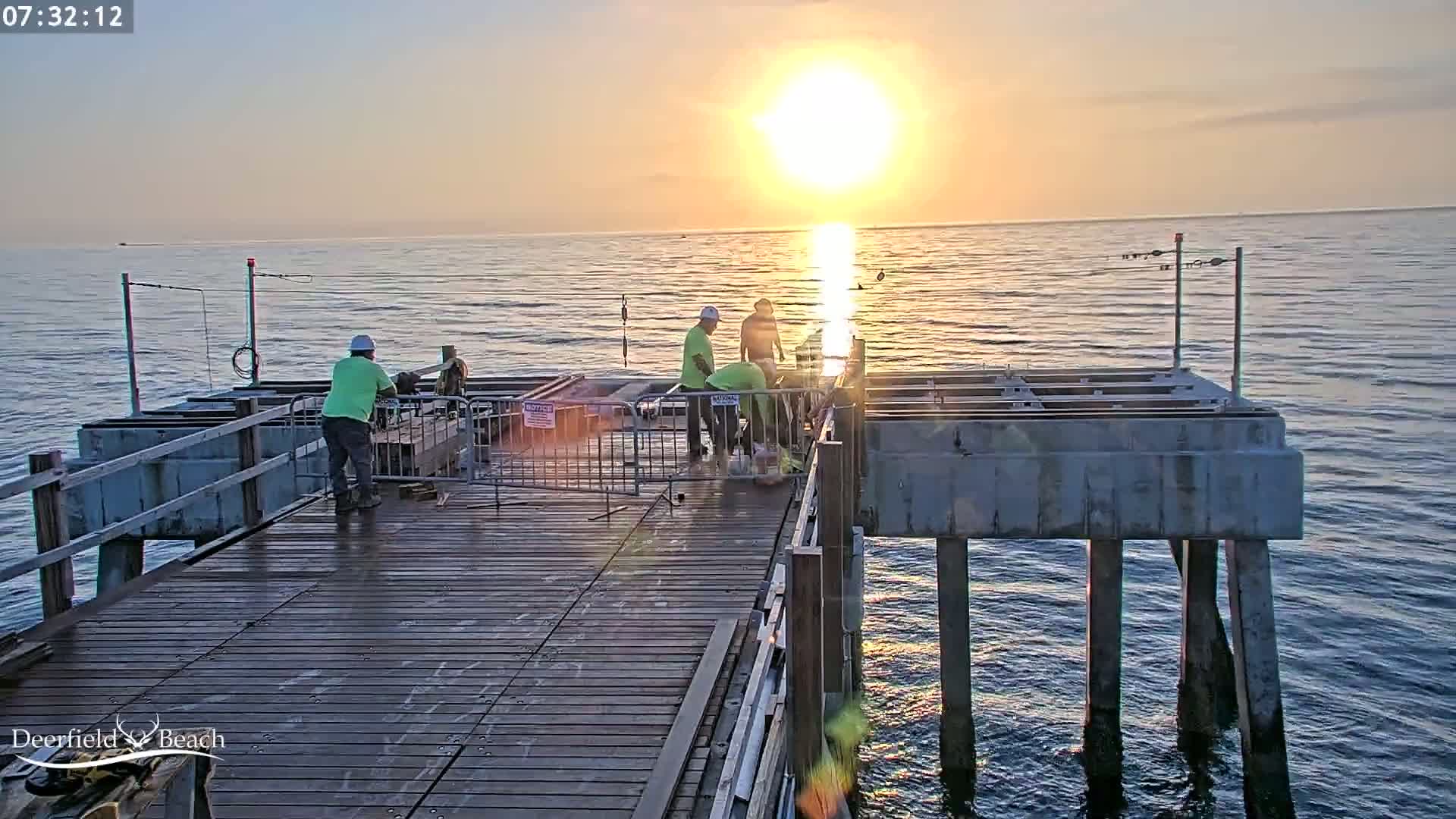 Several construction workers are working on a pier during a sunrise over calm ocean water.