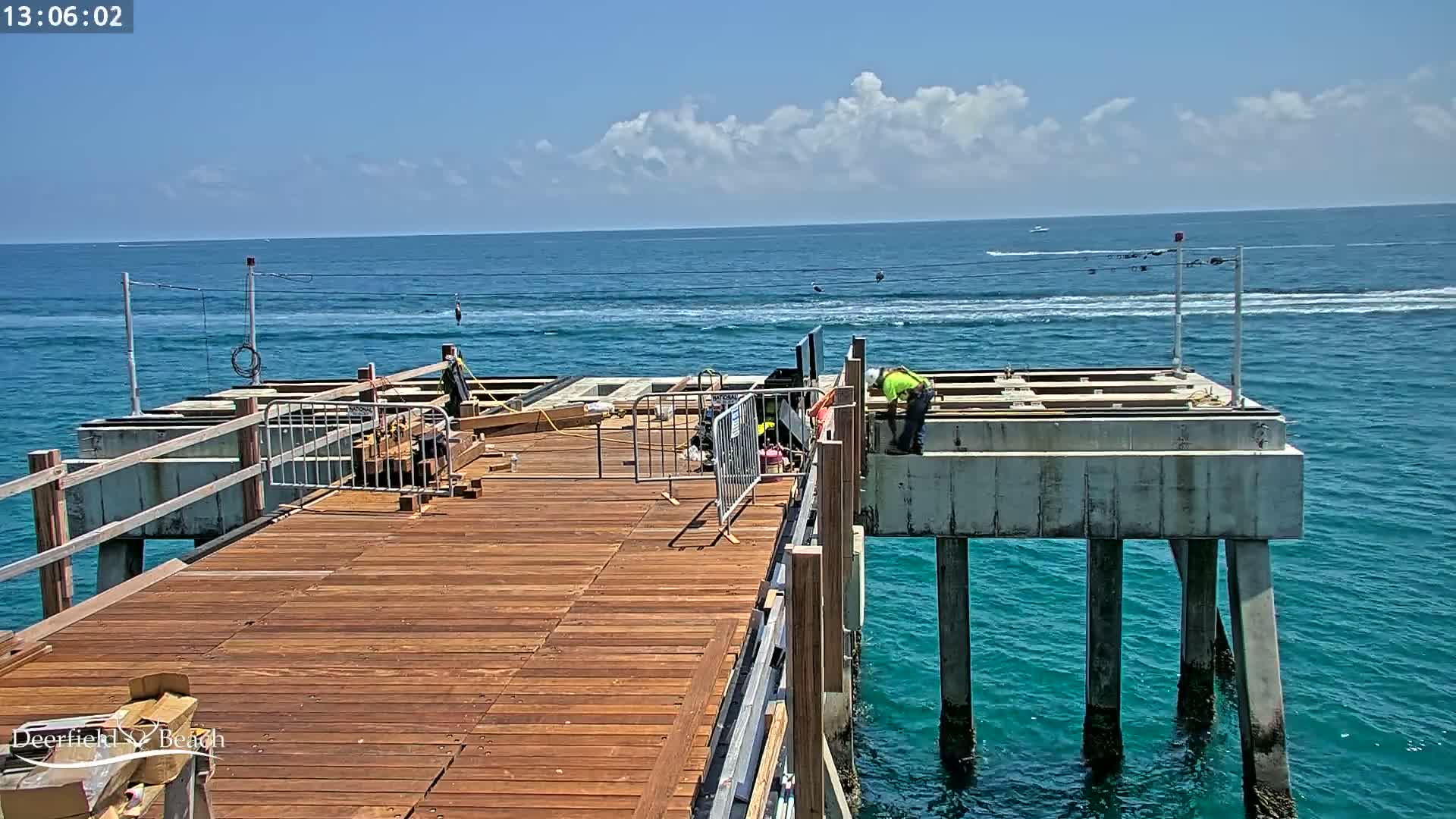 Construction workers are working on a wooden pier extending over a blue ocean under a partly cloudy sky.
