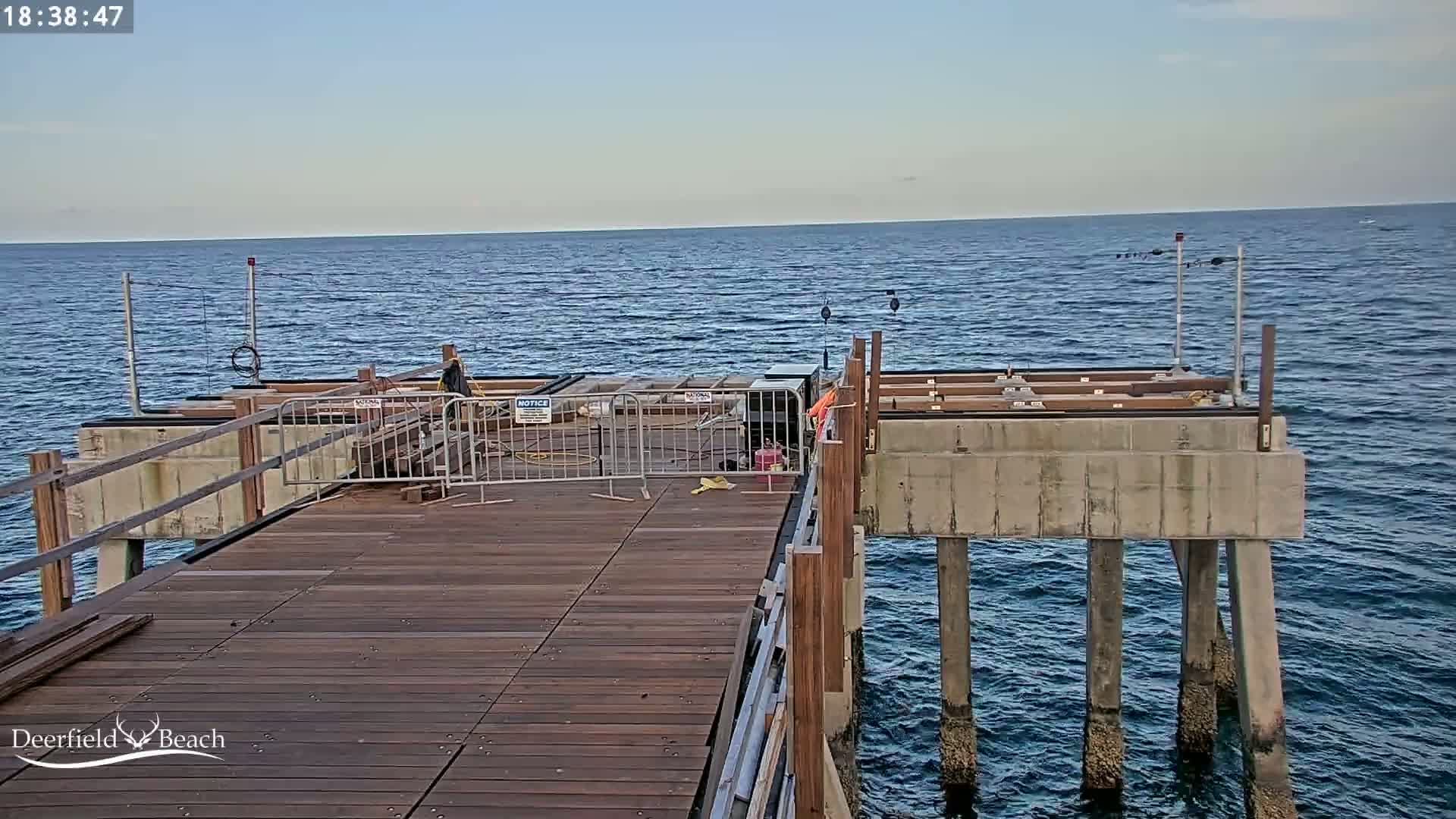 A partly constructed wooden pier extends over a calm ocean under a clear sky.
