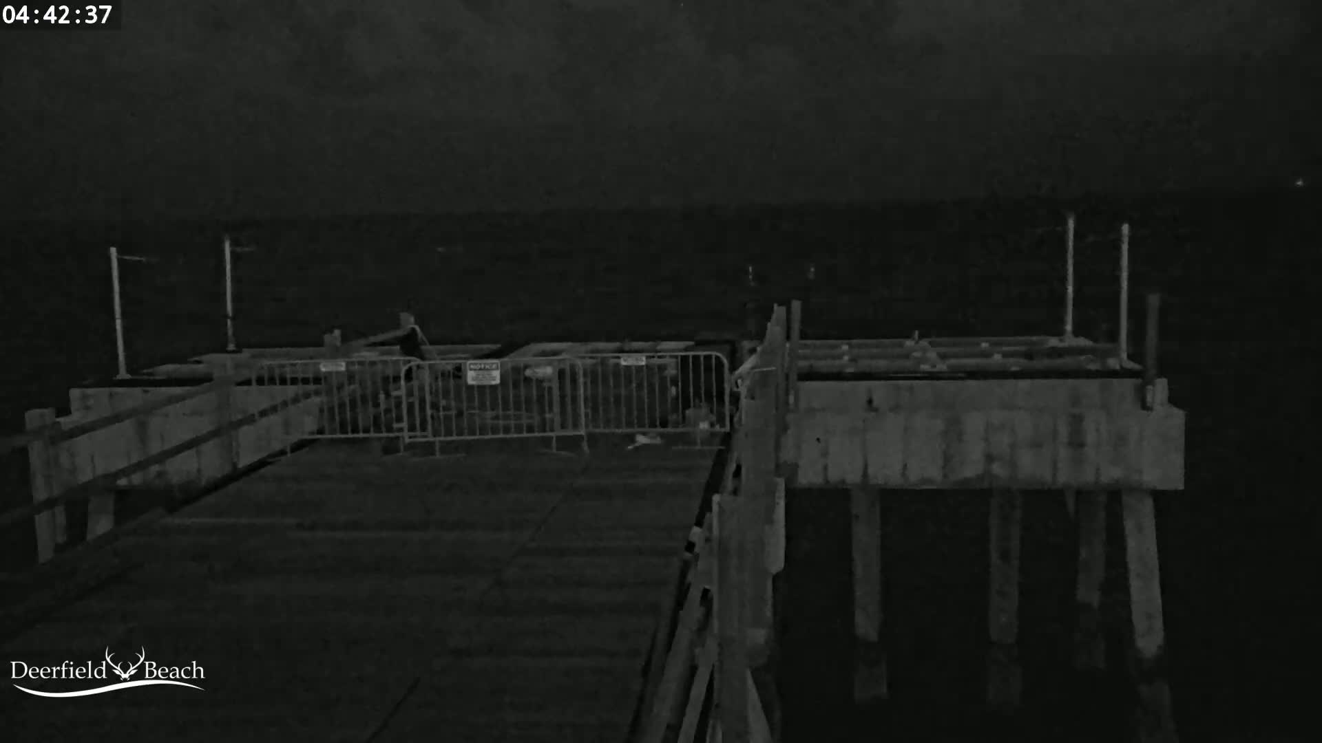 A nighttime, low-light image shows a section of a pier with temporary fencing and dark, possibly stormy, skies.