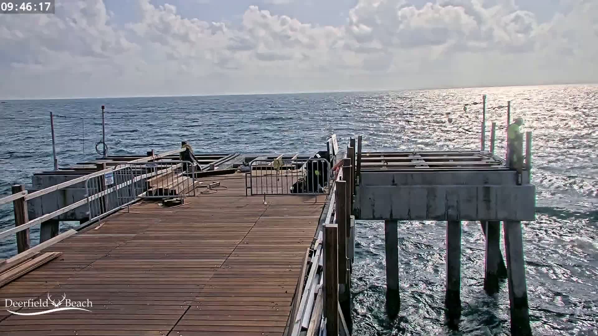 A partly sunny day shows construction workers on a wooden pier extending over a somewhat choppy ocean.