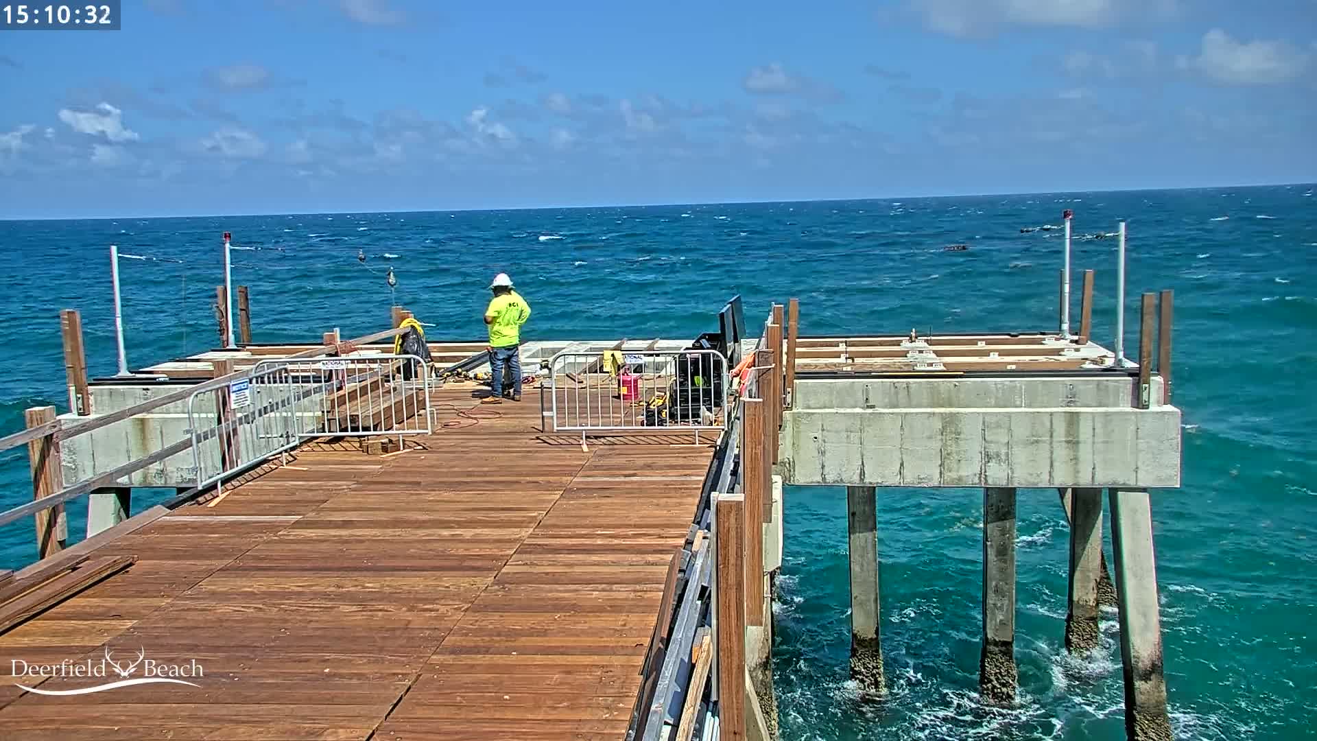 A partially-constructed pier extends over a teal ocean with moderate waves under a partly cloudy sky.