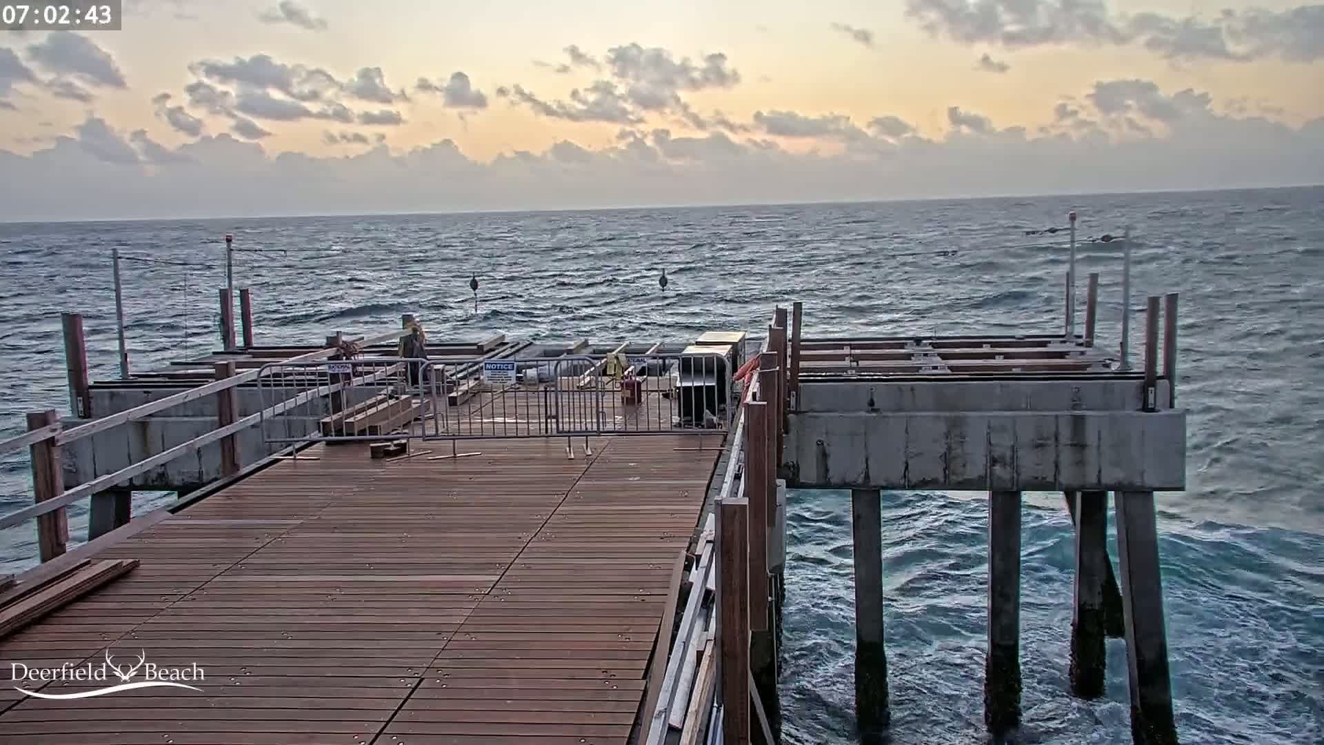 A partially constructed wooden pier extends over a wavy ocean under a partly cloudy, sunrise sky.
