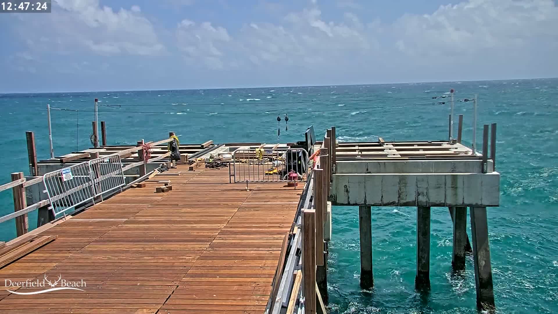 A partially constructed wooden pier extends over a teal ocean with small waves under a partly cloudy sky.
