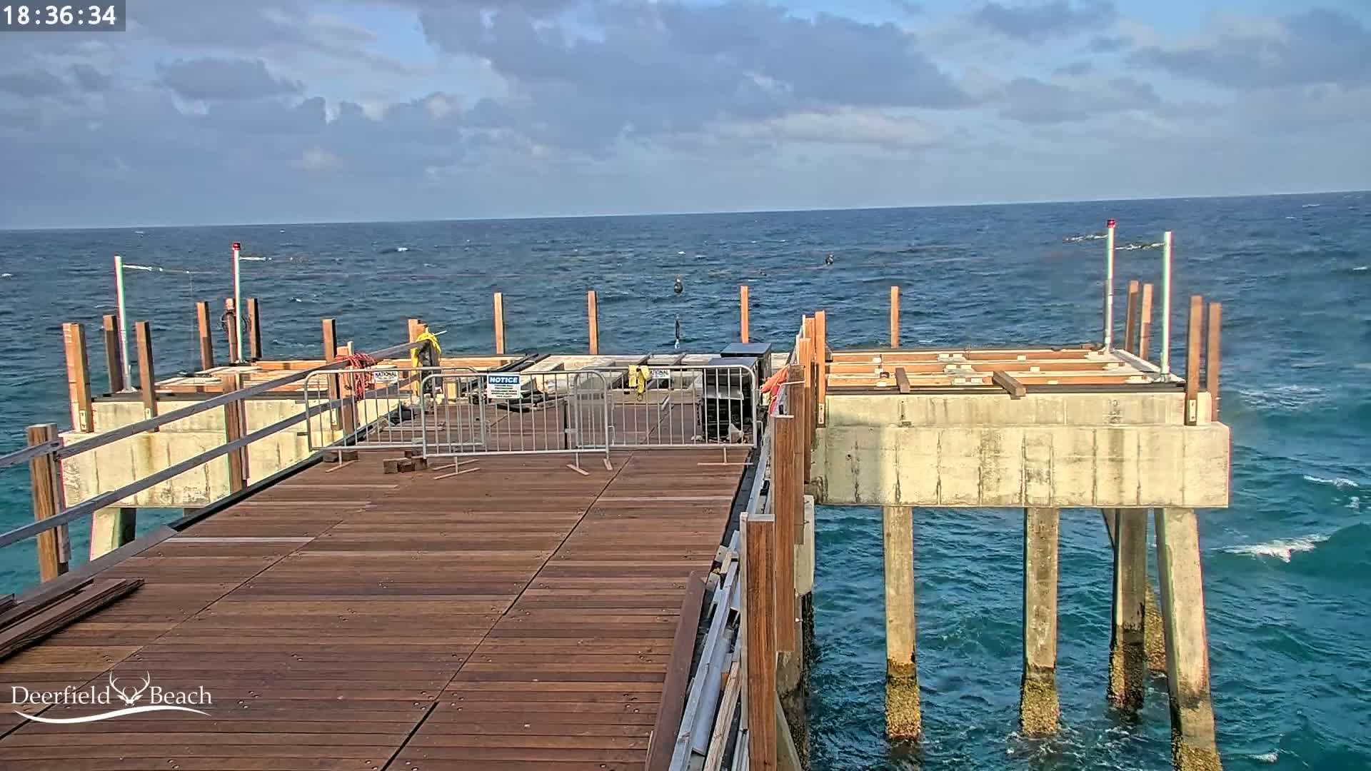 A partially constructed wooden pier extends over a blue ocean under a partly cloudy sky.
