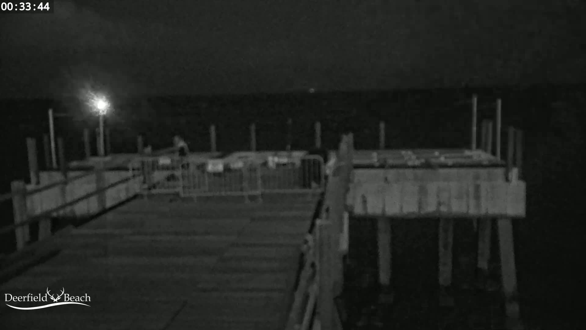 A nighttime, low-light image shows a mostly empty pier extending over dark water, with a single light source visible in the distance.