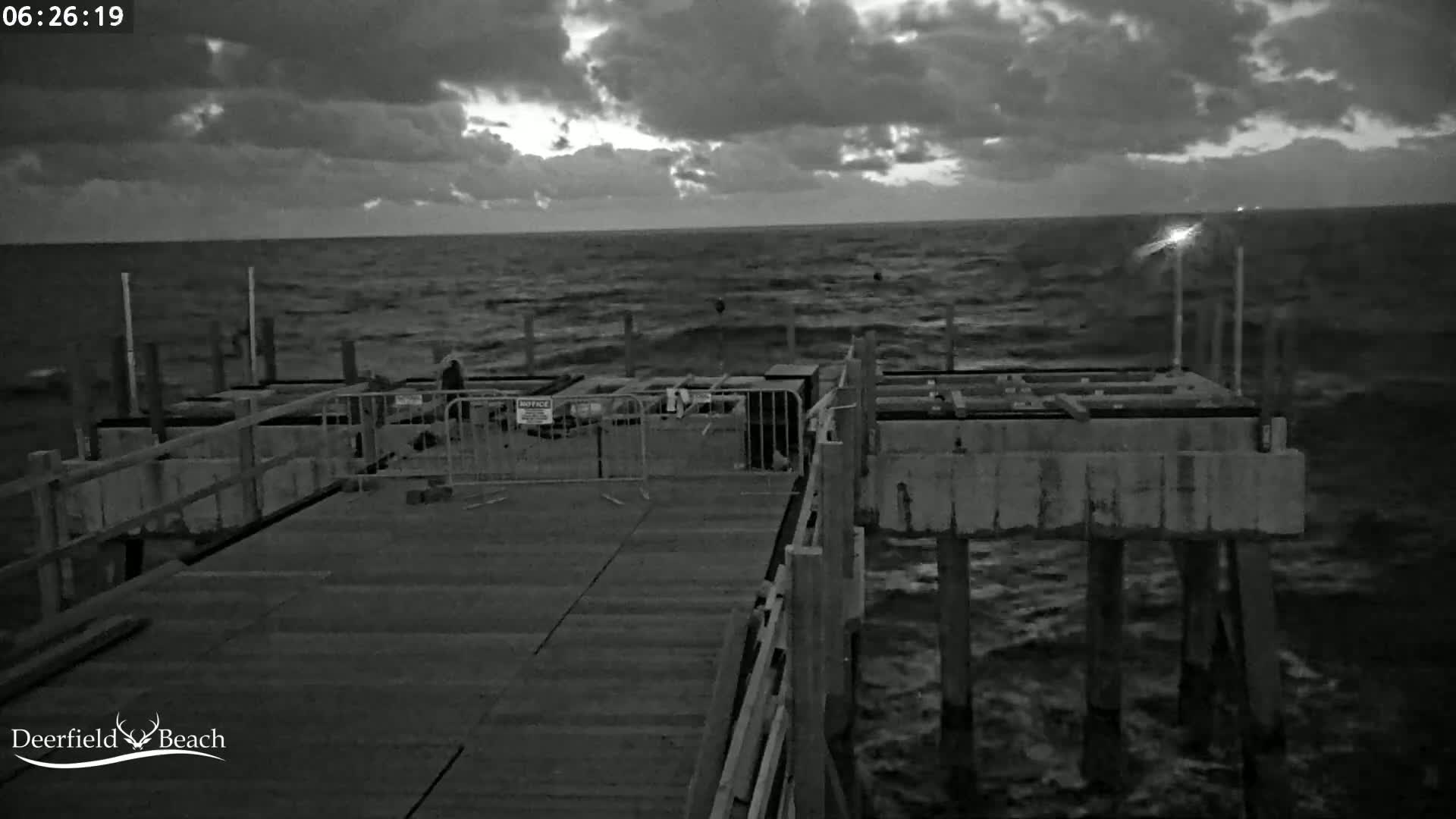 A partially constructed pier extends over a dark, choppy ocean under a cloudy, pre-dawn sky.