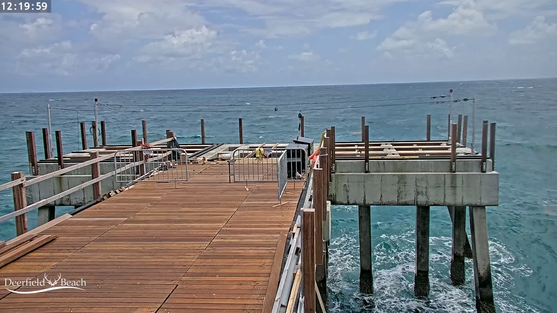 A partly-constructed wooden pier extends over a teal ocean with small waves under a partly cloudy sky.