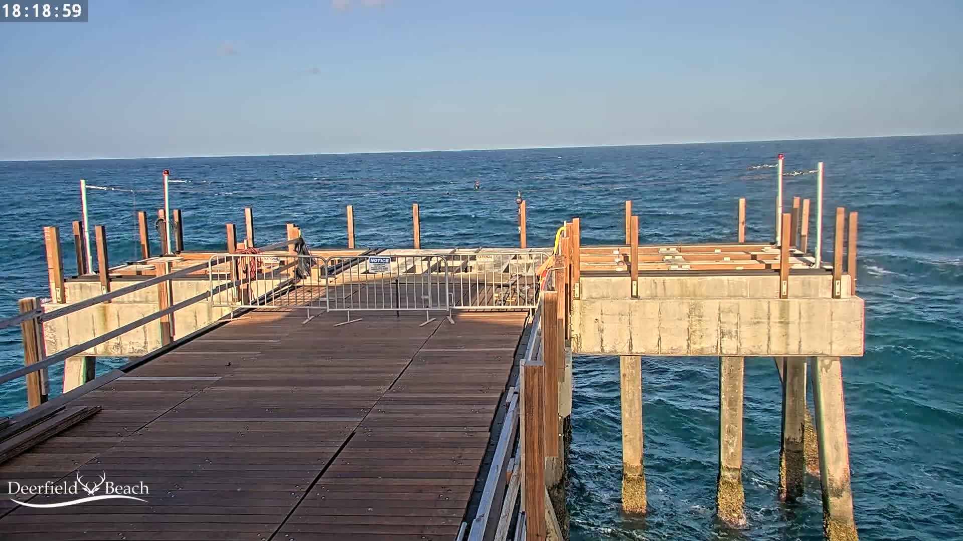 A partially constructed wooden pier extends over a blue ocean with small waves under a clear sky.