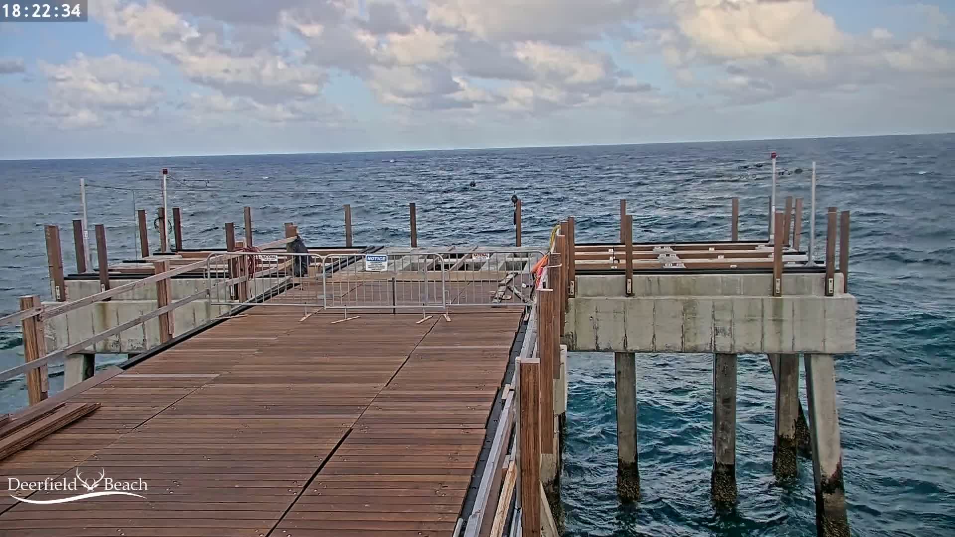 A partially constructed wooden pier extends over a dark blue ocean under a partly cloudy sky.