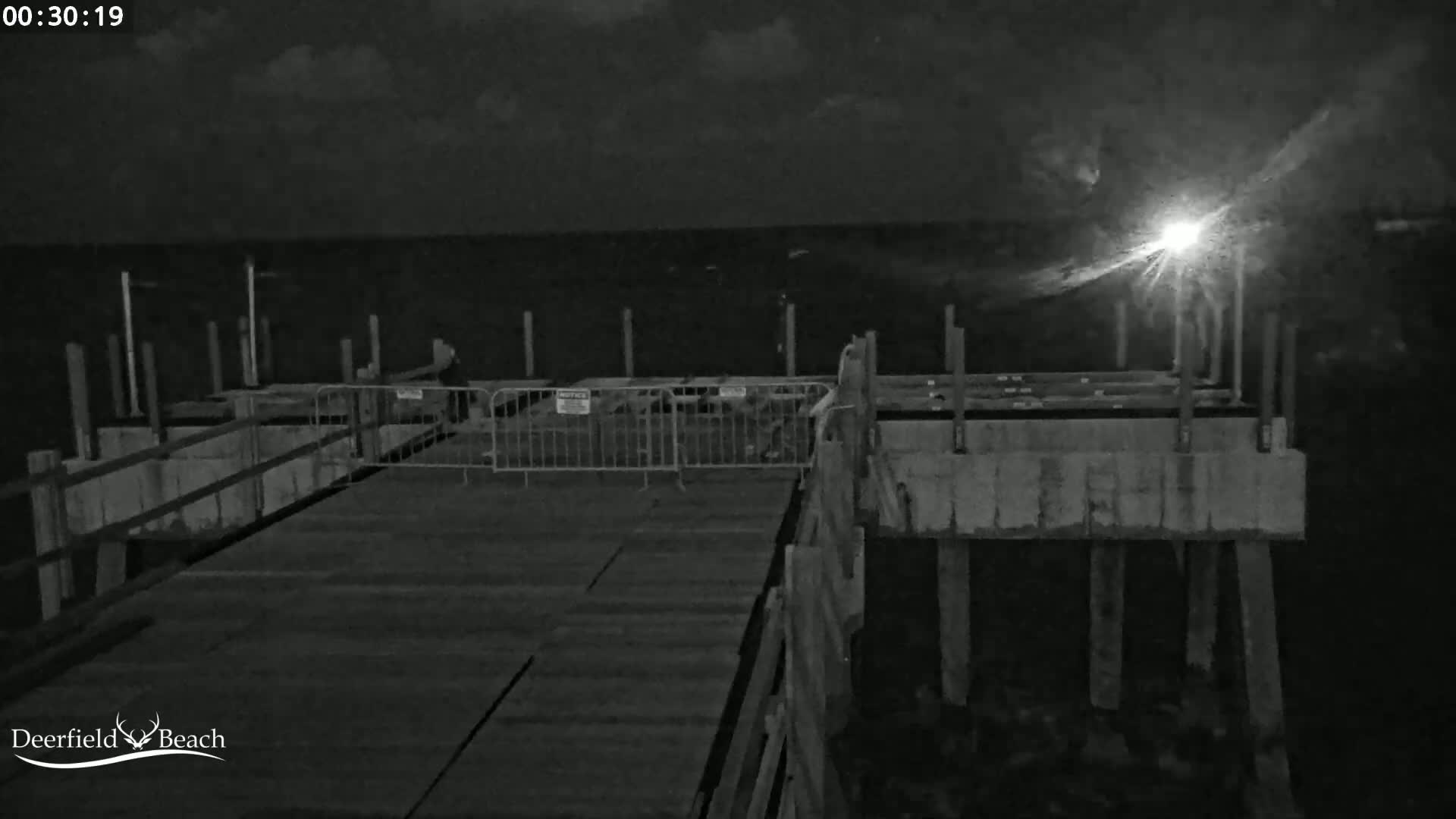 A nighttime view of a partially constructed pier extending over dark water under a mostly cloudy sky with a bright light source visible on the horizon.