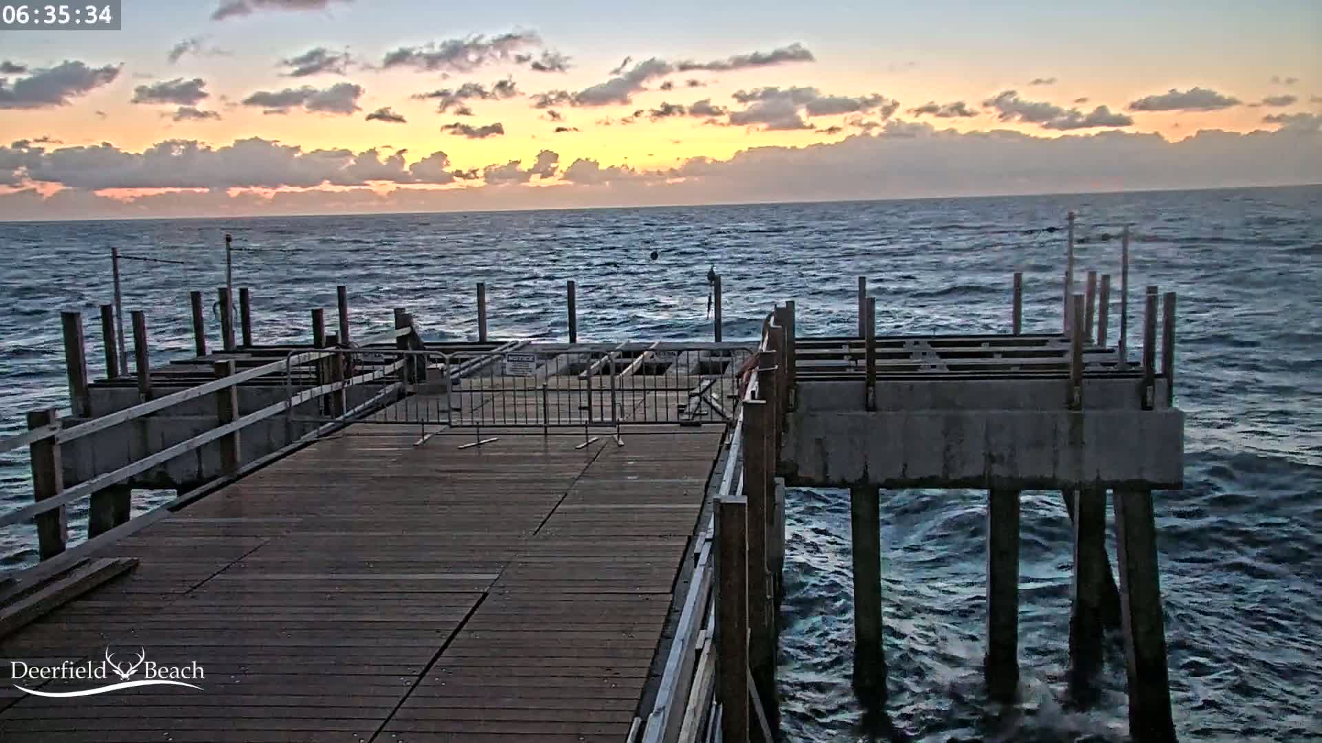 A wooden pier extends over a dark blue ocean during a sunrise with cloudy skies.