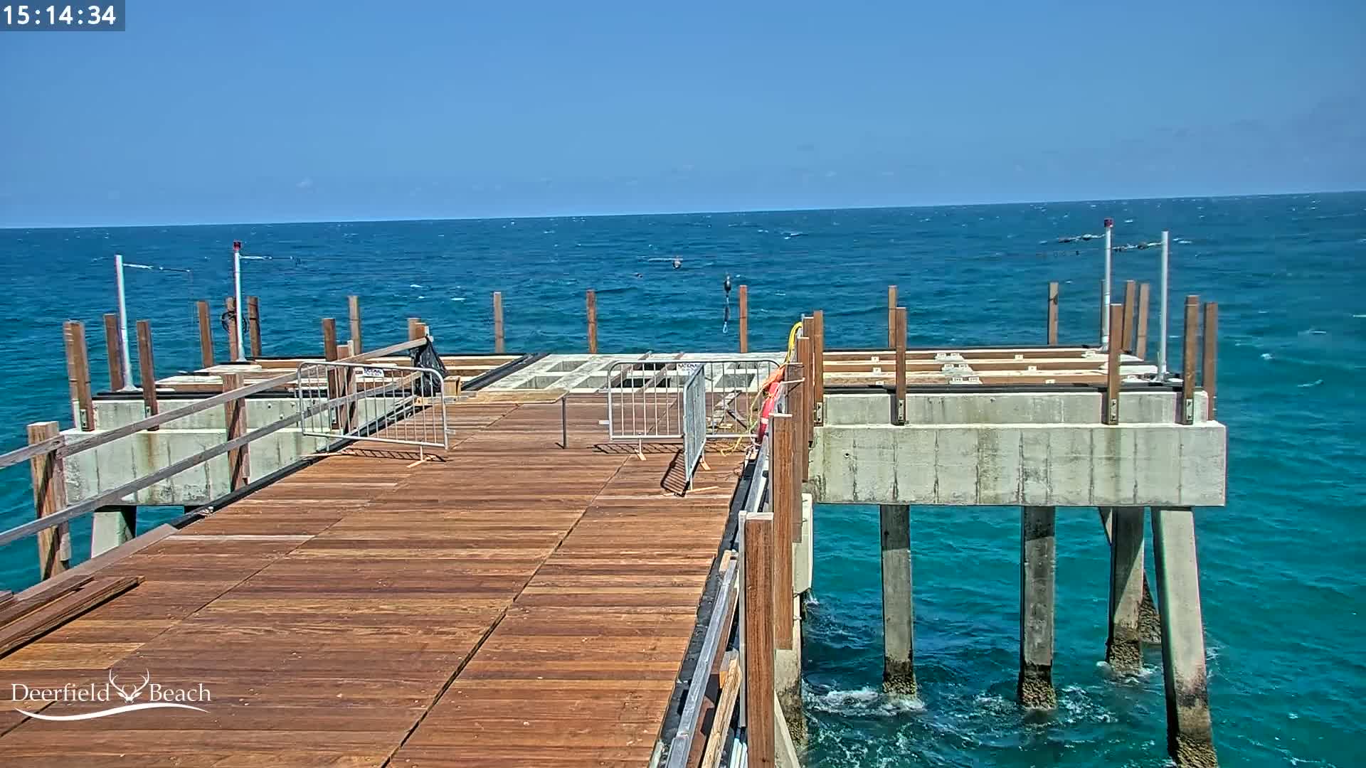 A partially constructed wooden pier extends over a bright blue ocean under a clear sky with light waves.