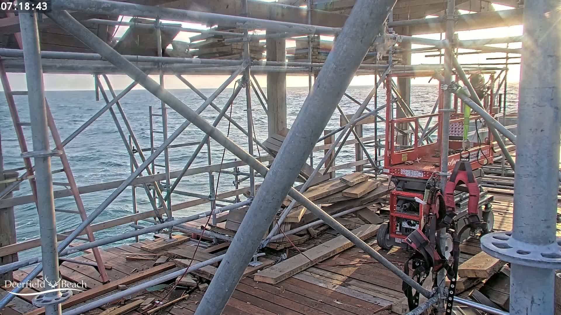 A partially constructed pier overlooking a wavy ocean under a mostly clear sky shows scaffolding, construction materials, and a red lift.