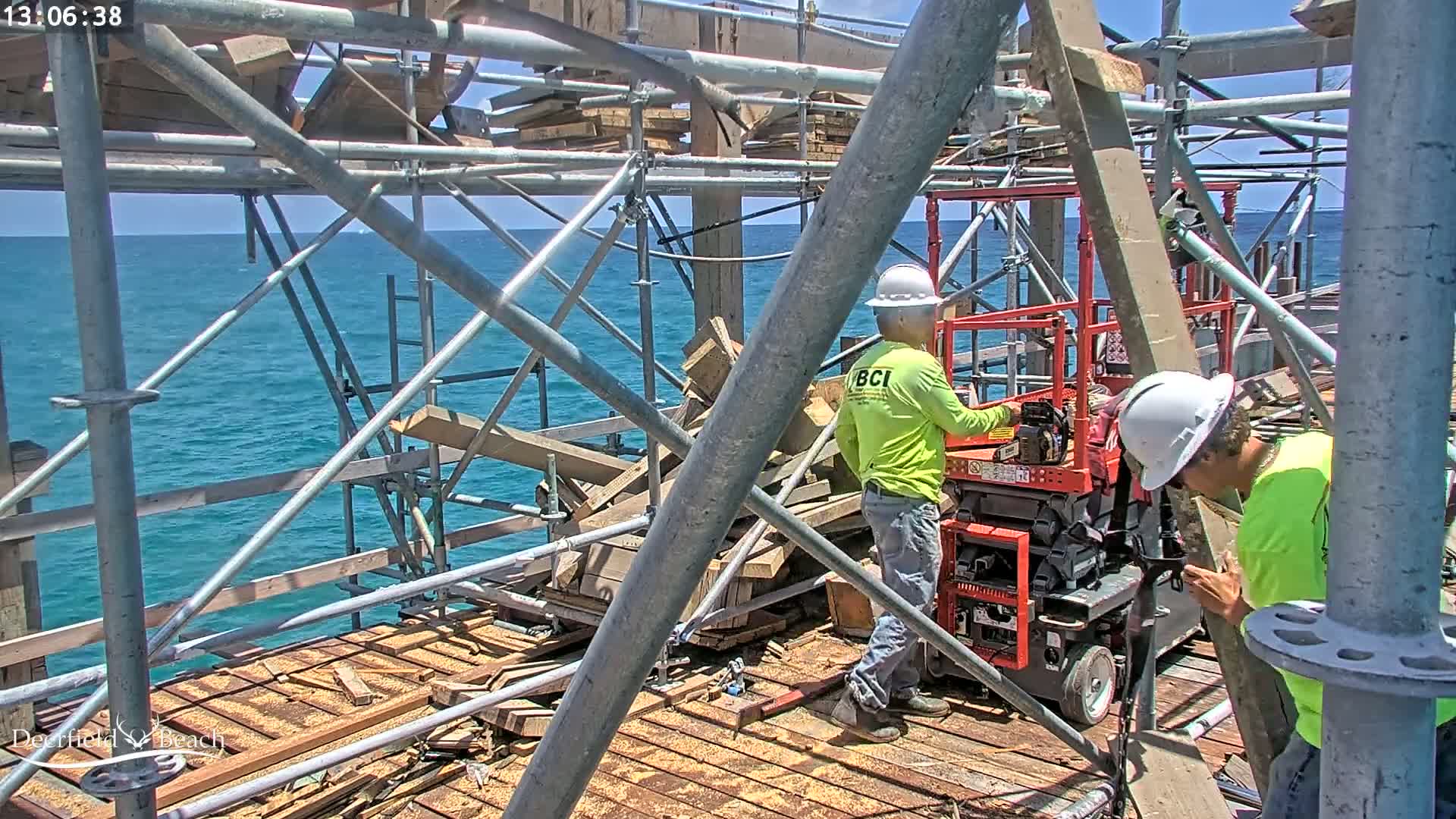 Two construction workers in hard hats and lime green shirts work on a scaffolding platform over a blue ocean on a sunny day.