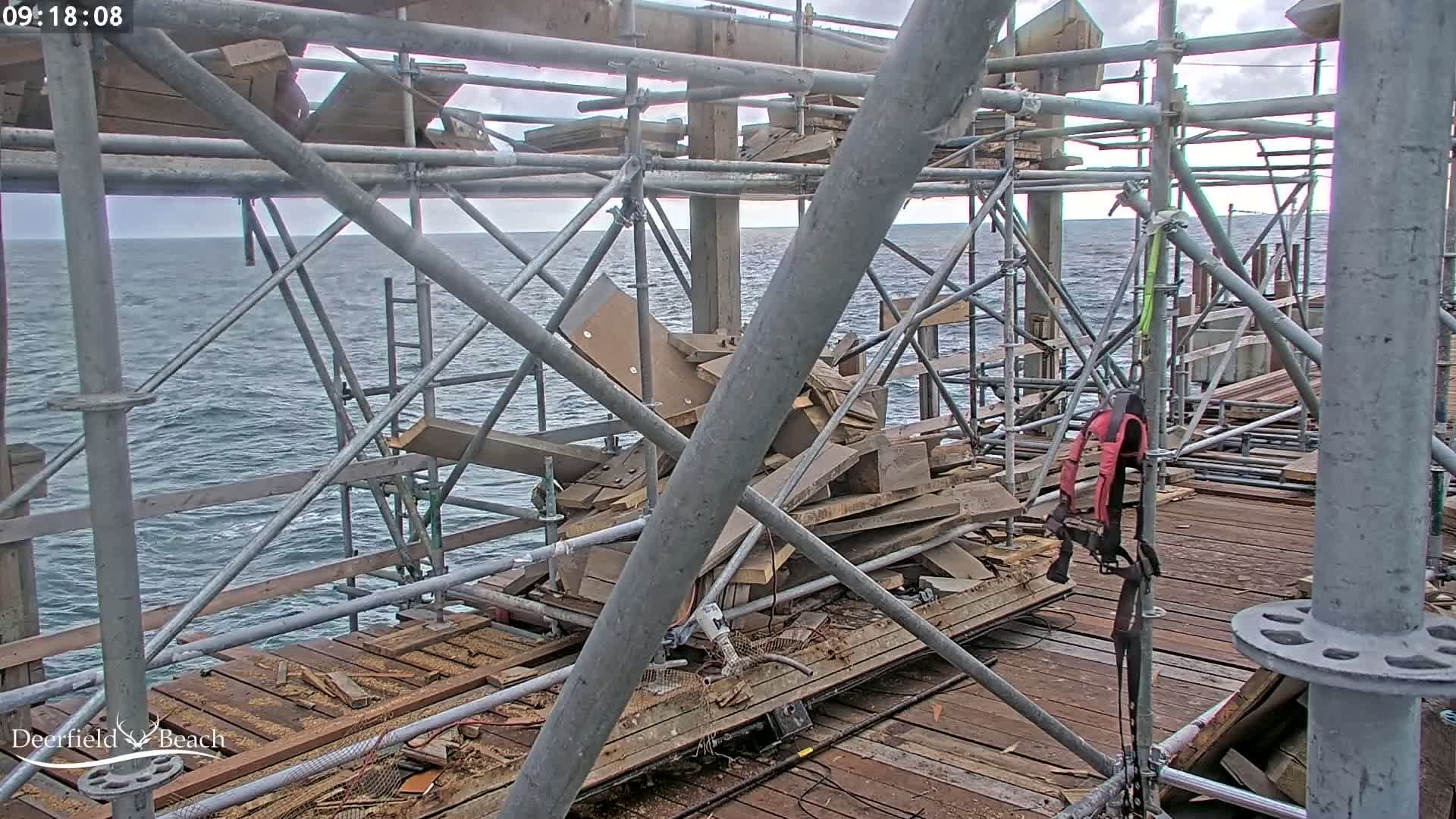 A partially constructed pier over a choppy sea under an overcast sky shows damaged wooden structures and scaffolding amidst debris.