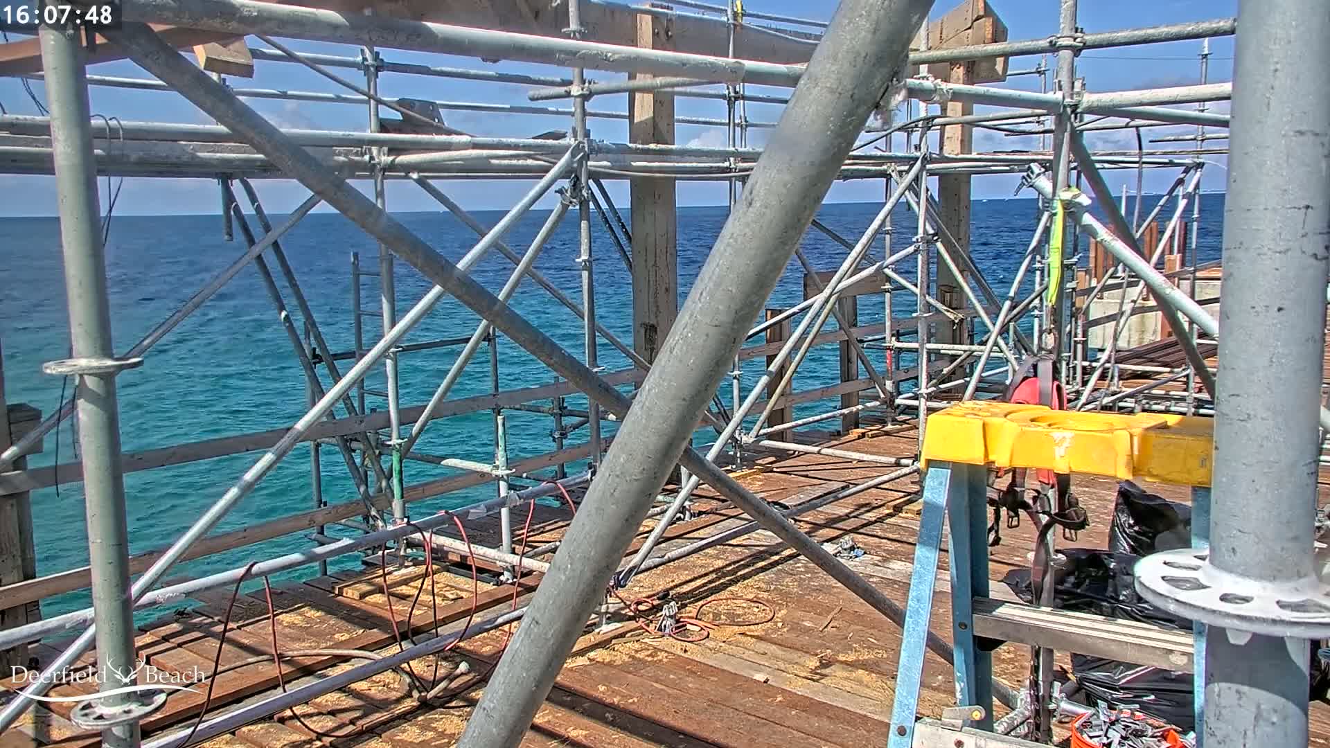 Metal scaffolding surrounds a wooden structure under construction overlooking a calm, blue ocean under a sunny sky.