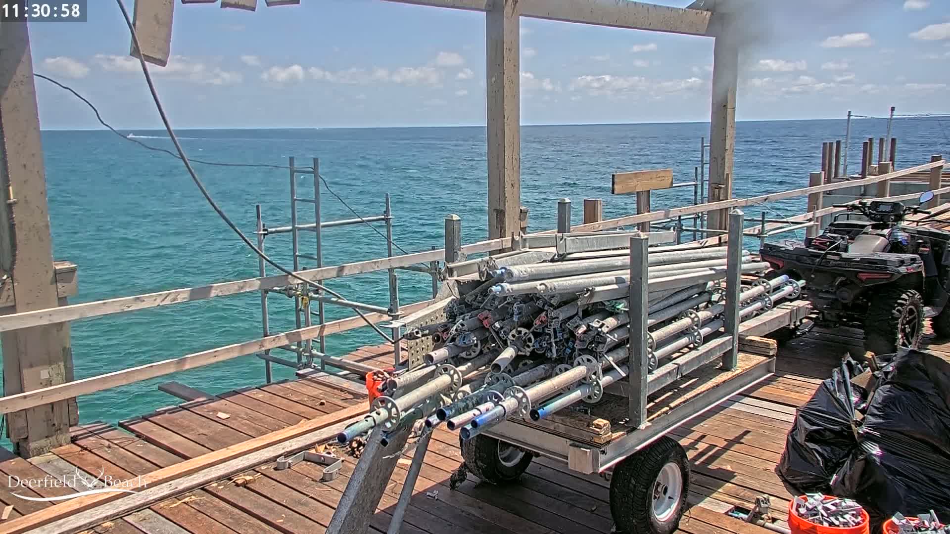 A flatbed trailer loaded with scaffolding is parked on a wooden pier overlooking a calm, blue ocean under a partly cloudy sky, with an ATV parked behind it.
