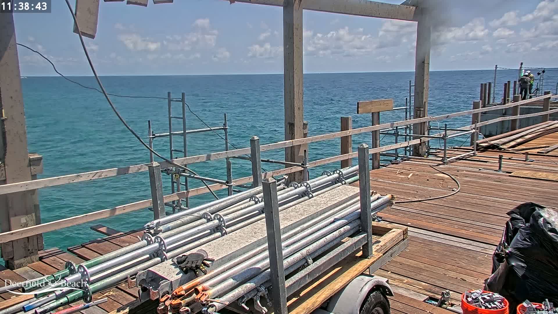 A partly cloudy day shows scaffolding and building materials on a wooden pier extending over a calm, teal ocean.