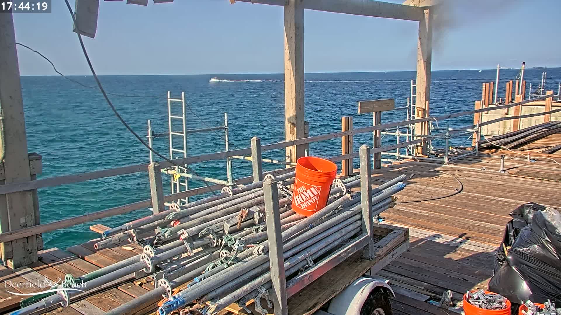 A sunny day shows a partially constructed wooden pier over blue ocean water; scaffolding and metal pipes are stacked on a cart near the pier's edge.