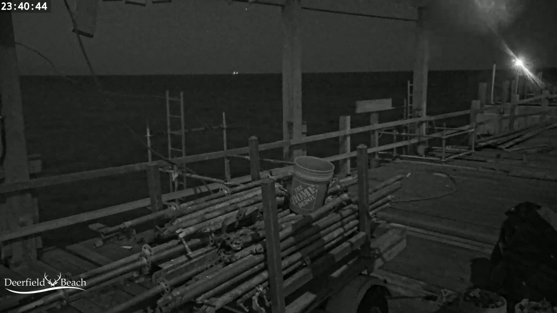 At night, a cart loaded with metal pipes and a bucket sits on a pier overlooking a dark body of water.